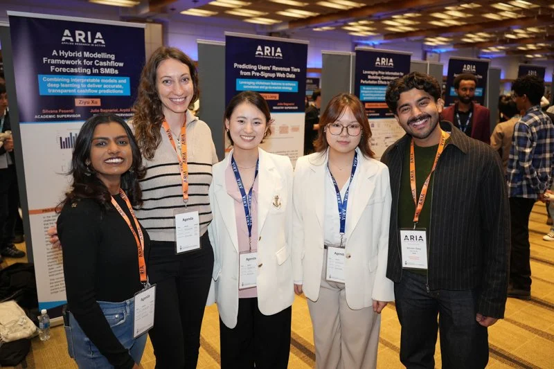  Five attendees wearing ARIA lanyards and badges standing together in front of research posters at the ARIA event exhibit area. 
