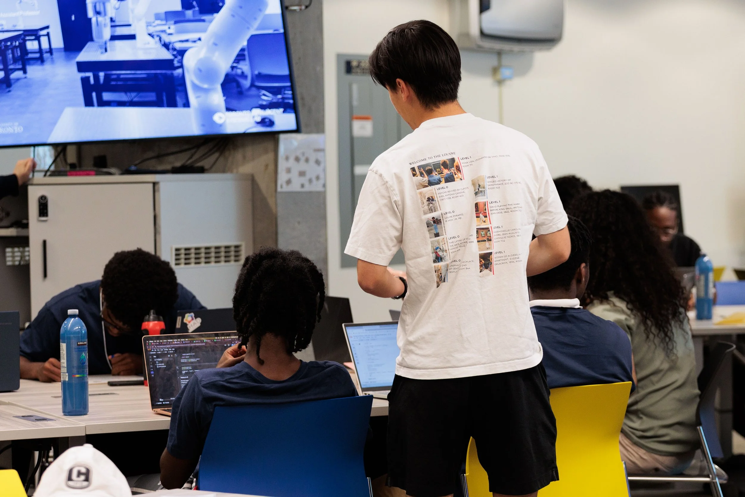 Group of participants working on laptops around tables in a classroom with a mentor standing behind looking over.