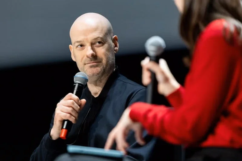 Close-up of a person holding a microphone on stage, looking toward another speaker in the foreground during a live discussion.