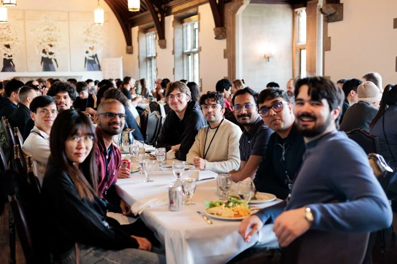 Graduate visitors sit at a long dining table during a group meal, with plates of food, glasses, and laptops visible in a historic hall with tall windows and wooden beams.