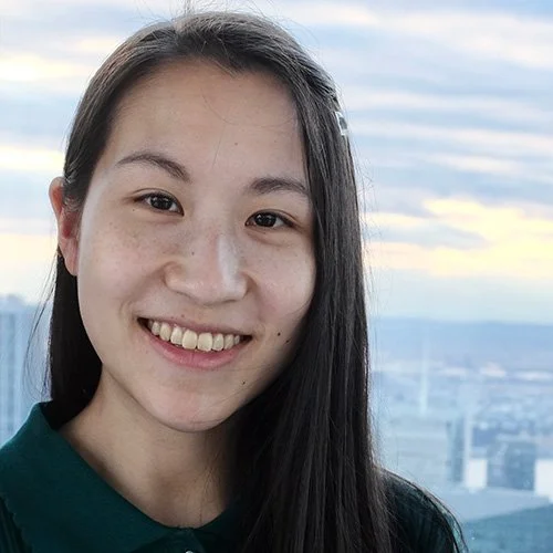Michelle Lam smiles facing the camera. Buildings are visible in the background.