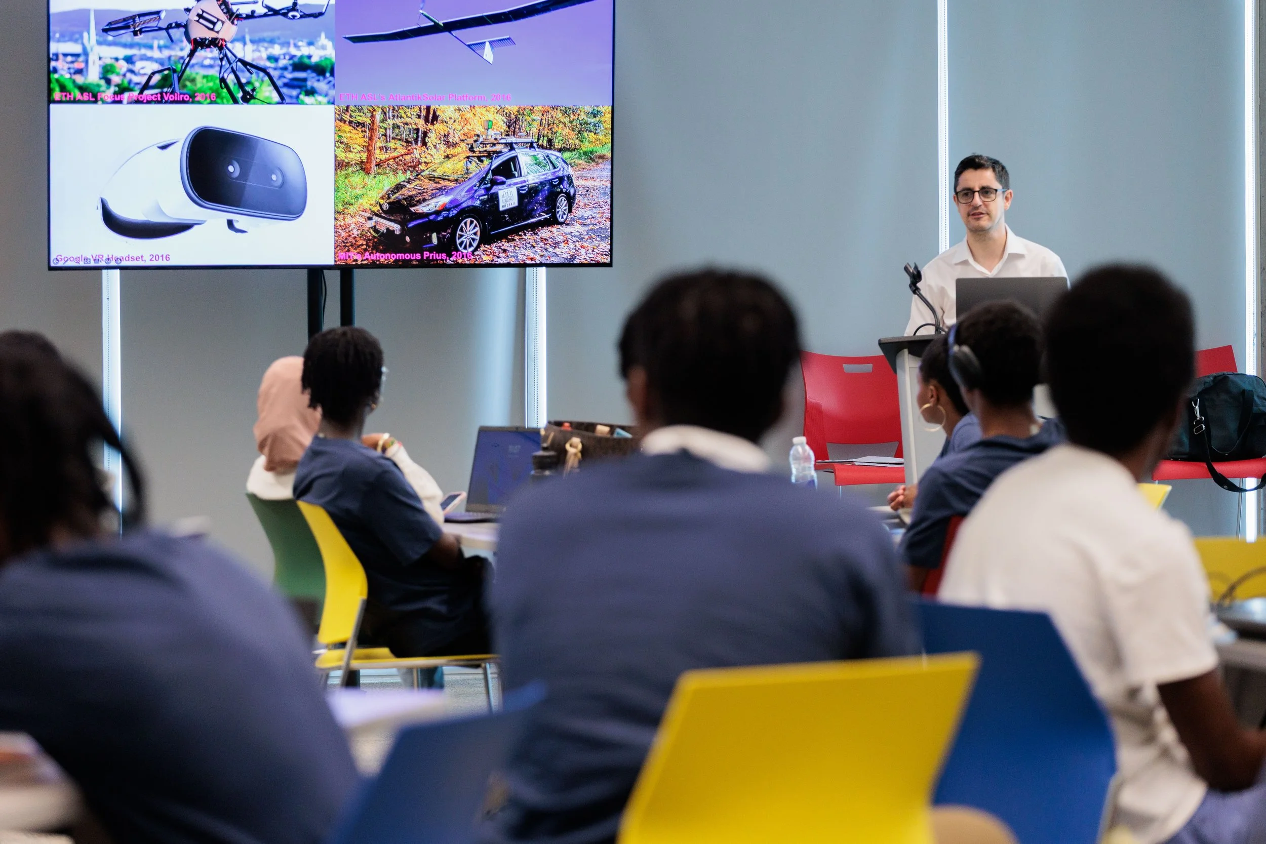 Presenter giving a presentation from a podium while attendees watch from their seats. A large display screen shows images of emerging technologies, including a drone, a virtual reality headset, and an autonomous vehicle.