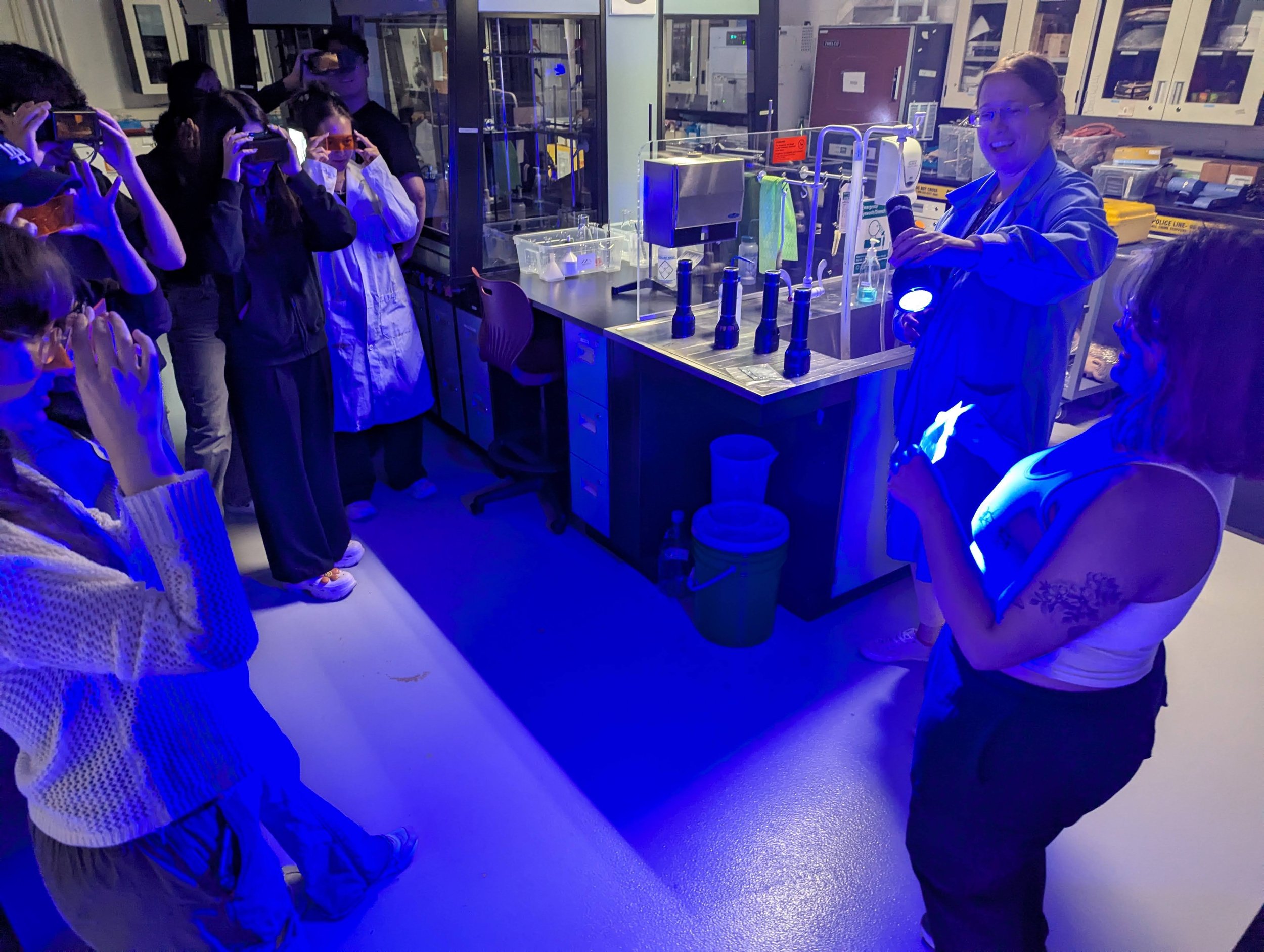 One student and a lab tech demonstrate a fingerprint with UV light while a group of students view the fingerprint through orange glasses.