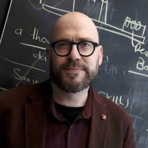 Alán Aspuru-Guzik stands in front of a chalkboard while smiling and facing the camera.