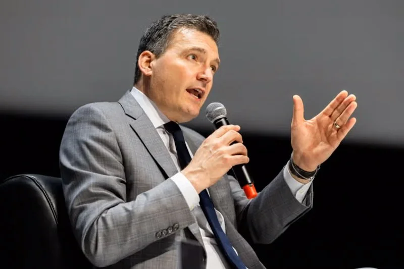 Person in a grey suit speaks into a handheld microphone on stage, gesturing with one hand during a panel discussion.