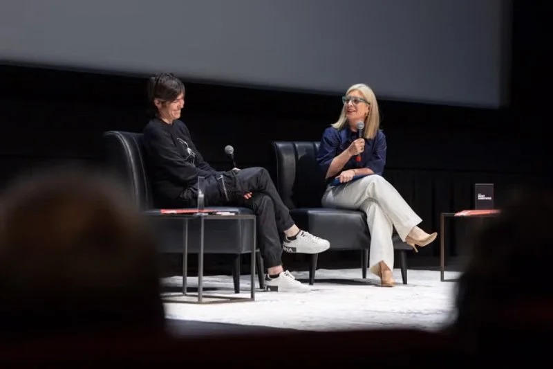 Two people seated on stage in armchairs, each holding a microphone and speaking during a live discussion, with a small table between them and a large screen in the background.