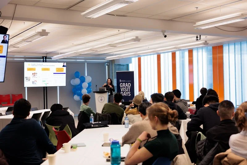 A speaker presents to a seated audience during Grad Visit Days in a classroom, with a podium, presentation screen, and event signage visible.