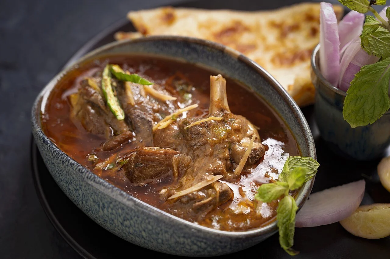 A steaming bowl of Mutton Nihari garnished with ginger julienne and green chilies, served with a side of naan bread, sliced red onions, and fresh mint leaves.
