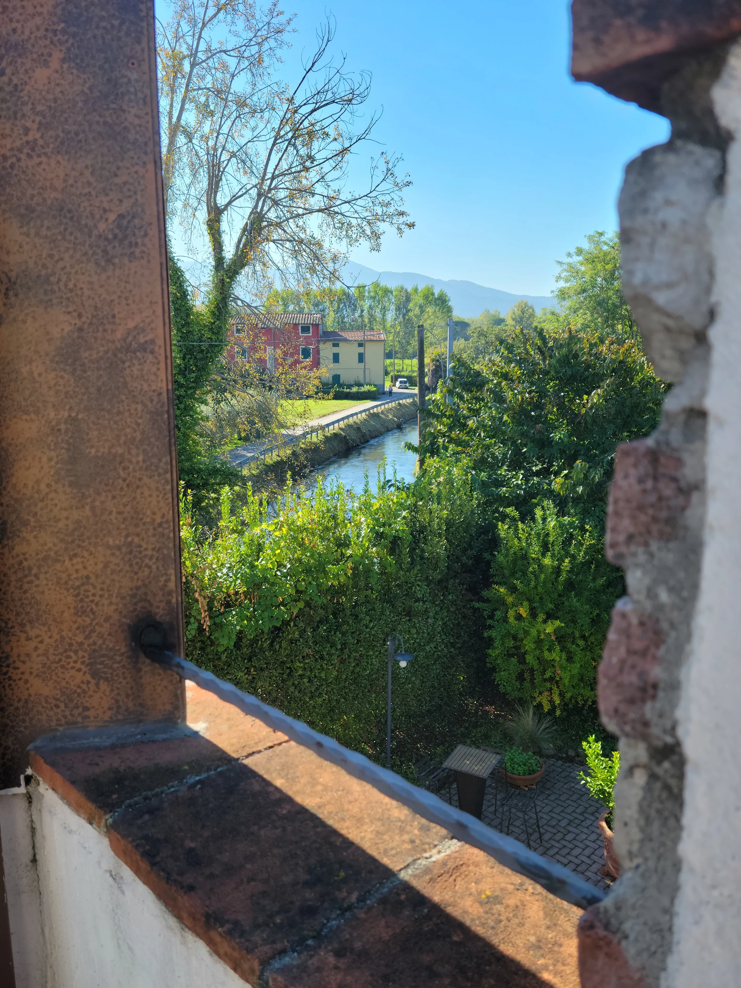A view from a stone castle balconette overlooking a small canal, lush green gardens, and a rustic red farmhouse in the Lucca countryside.