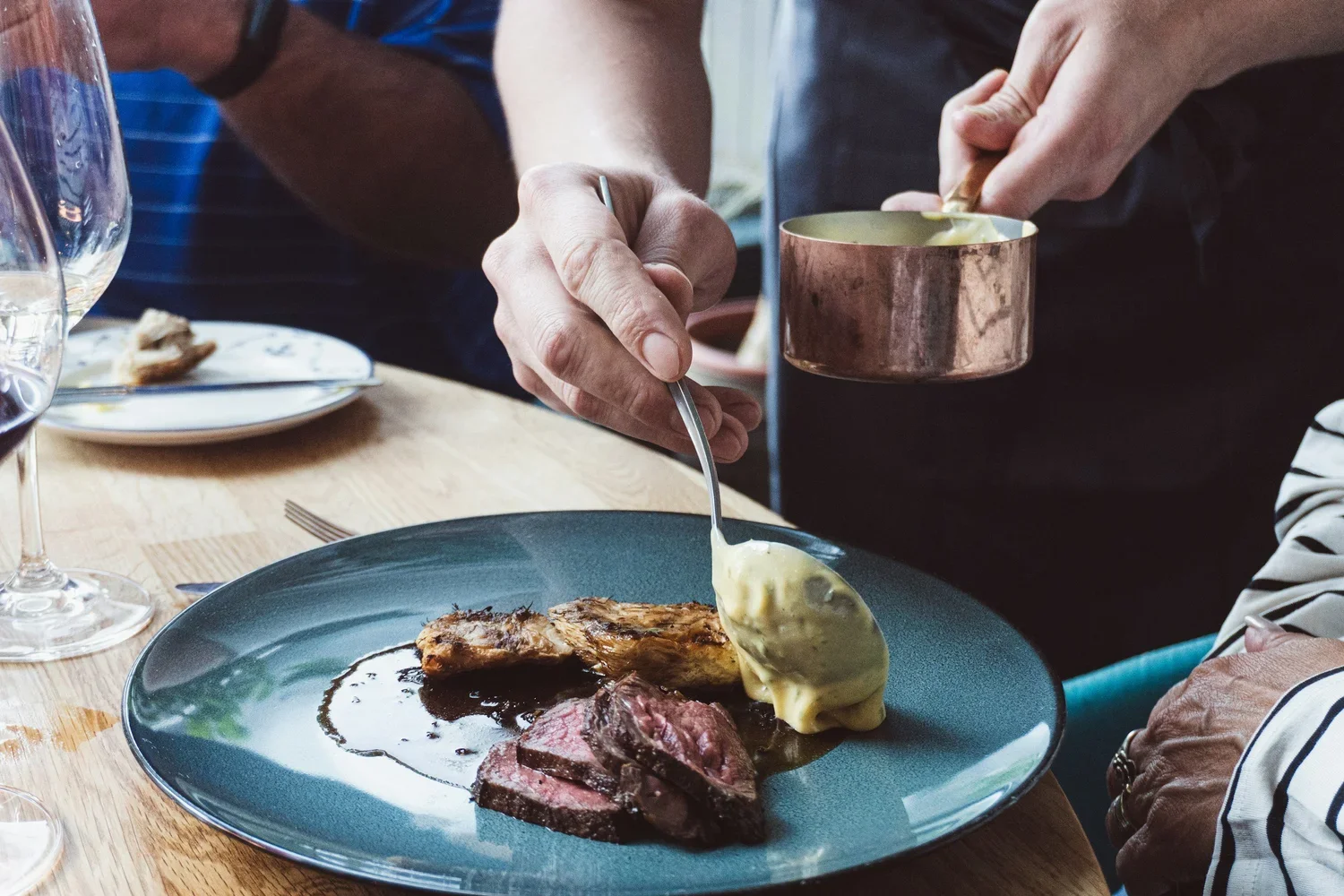 A chef using a spoon to pour creamy yellow hollandaise sauce over sliced medium-rare steak and roasted potatoes on a blue plate.