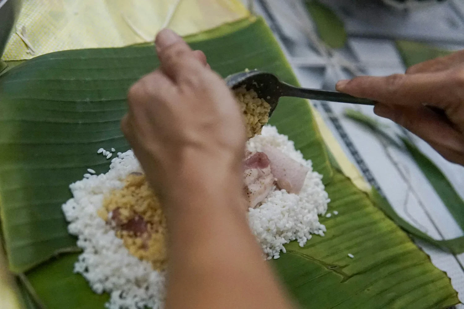 Hands layering mung beans and pork over glutinous rice on green dong leaves to make traditional Bánh Chưng for Tet in Hanoi.