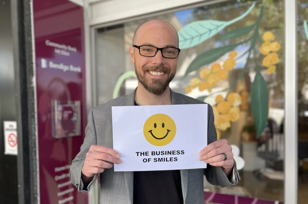 A smiling young man with glasses holding a sign that says The Business of Smiles, standing outside a bank