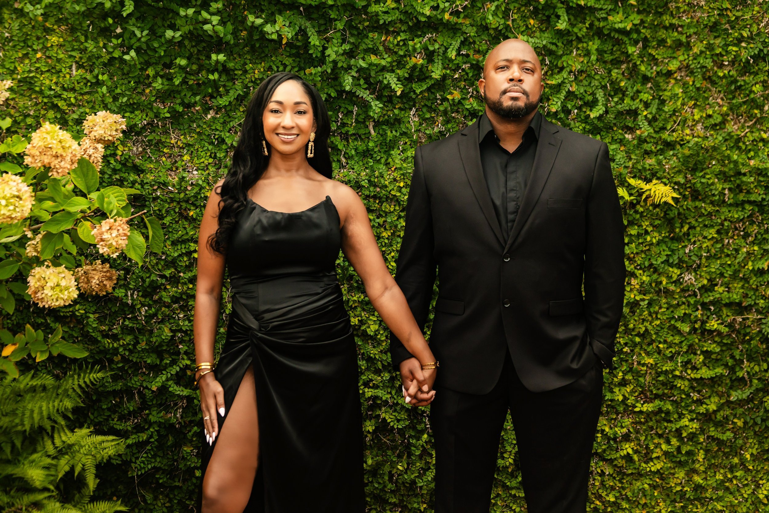 Couple holding hands wearing formal black attire standing against a lush green ivy wall with hydrangea flowers.