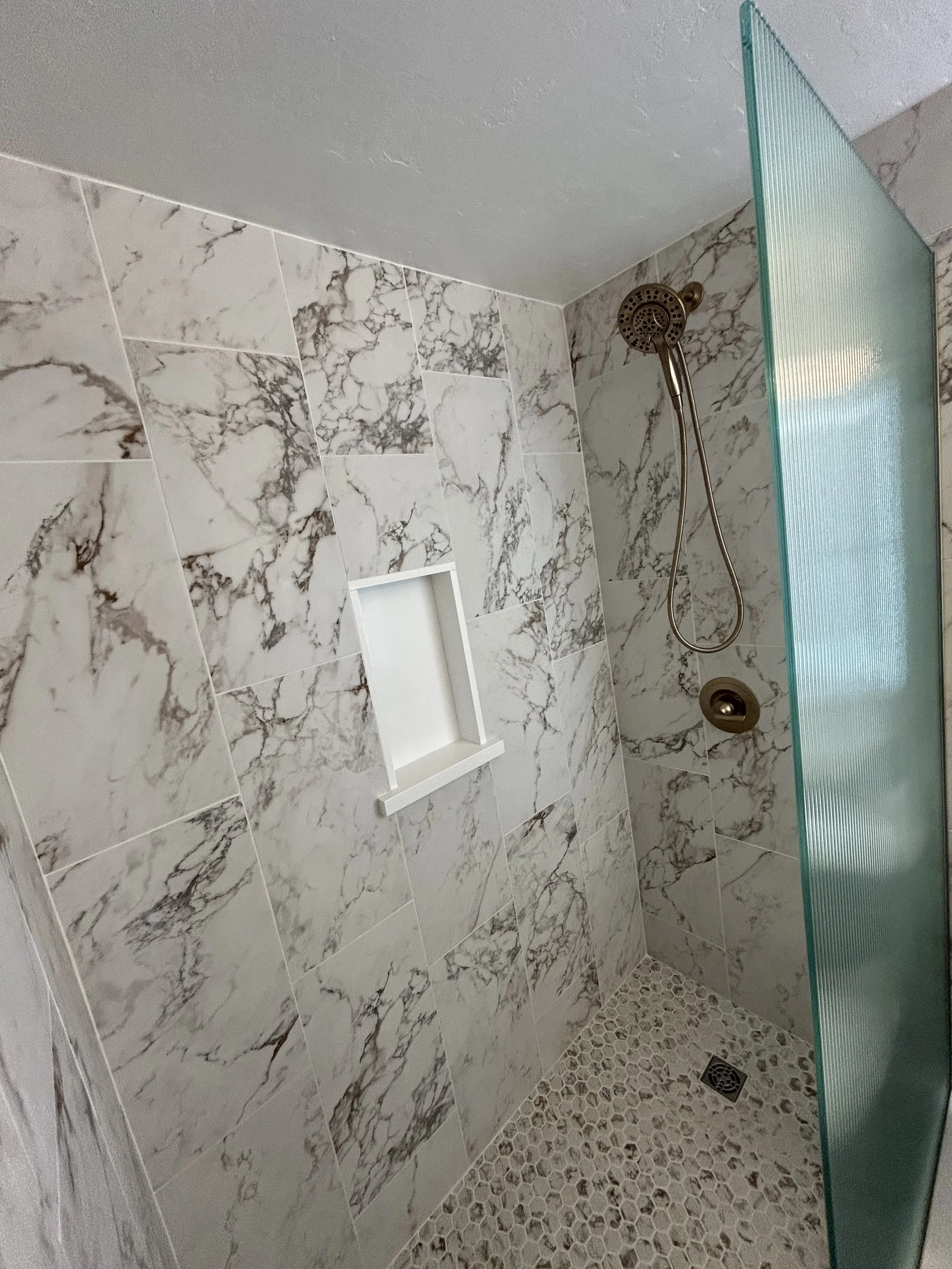 A modern walk-in shower with marble tile walls, a glass door, a showerhead, a built-in niche, and pebble floor tiles.