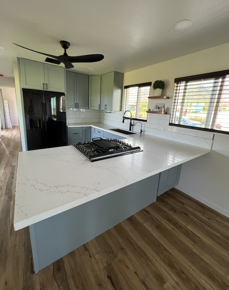 Modern kitchen with blue cabinets, white marble countertop, black refrigerator, black stove, and wooden flooring. Large windows with blinds, black ceiling fan, and small decorative shelves with plants and photos.