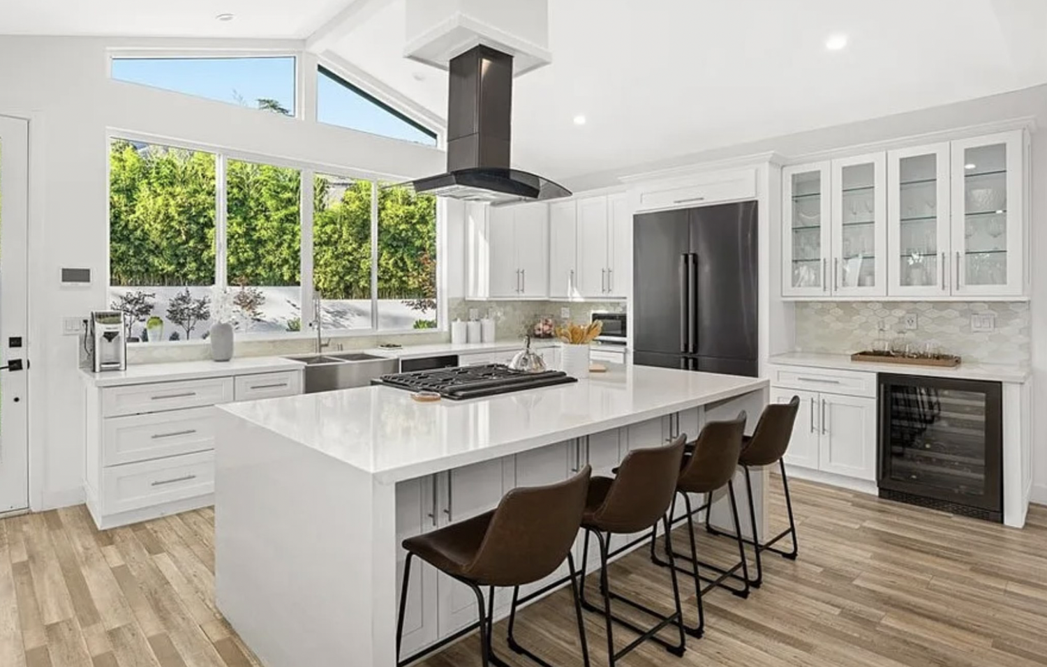 Modern white kitchen with large island, brown chairs, stainless steel appliances, wood flooring, and large windows showing greenery outside.