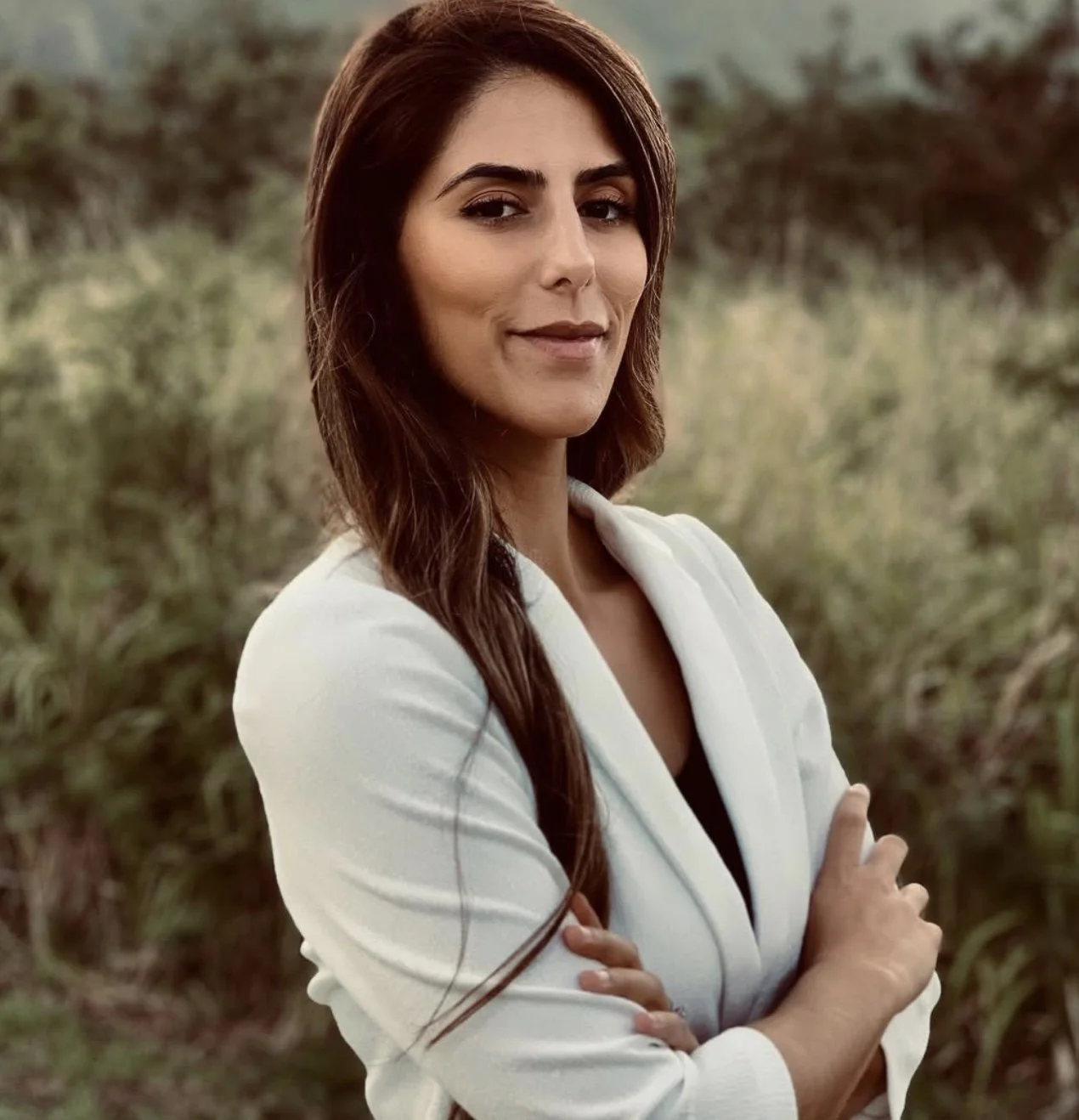 A woman with long brown hair, wearing a white blazer, standing outdoors with arms crossed and a slight smile, with green foliage in the background.