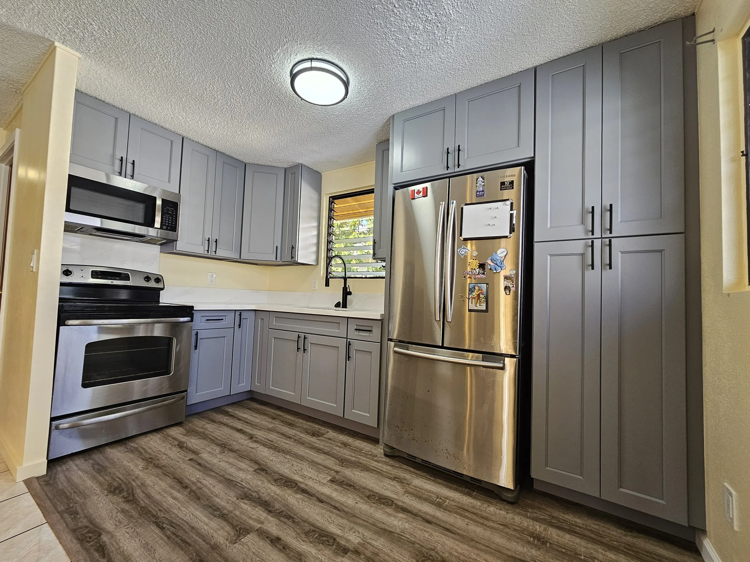 Modern kitchen with gray cabinets, stainless steel appliances including a fridge, oven, microwave, and a black sink with a window above, wooden floor, ceiling light fixture.