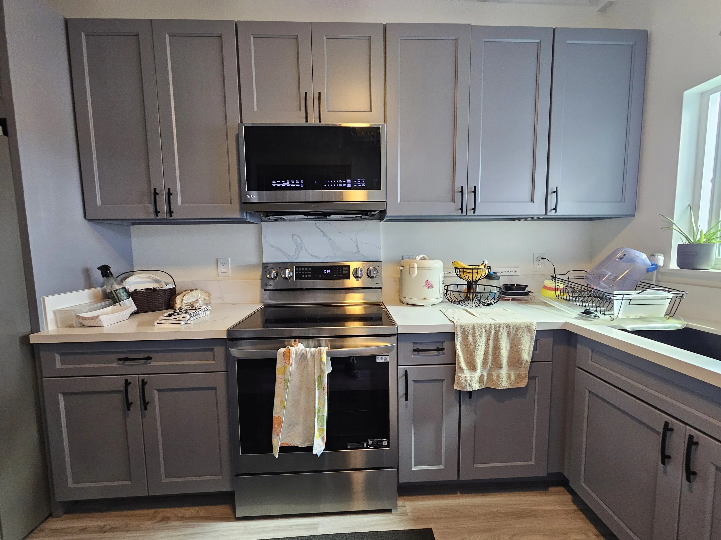Kitchen with gray cabinets, stainless steel stove, microwave above, white countertops, dish drying rack with dishes, dish soap, towel, electric rice cooker, hand soap, and window with potted plant.