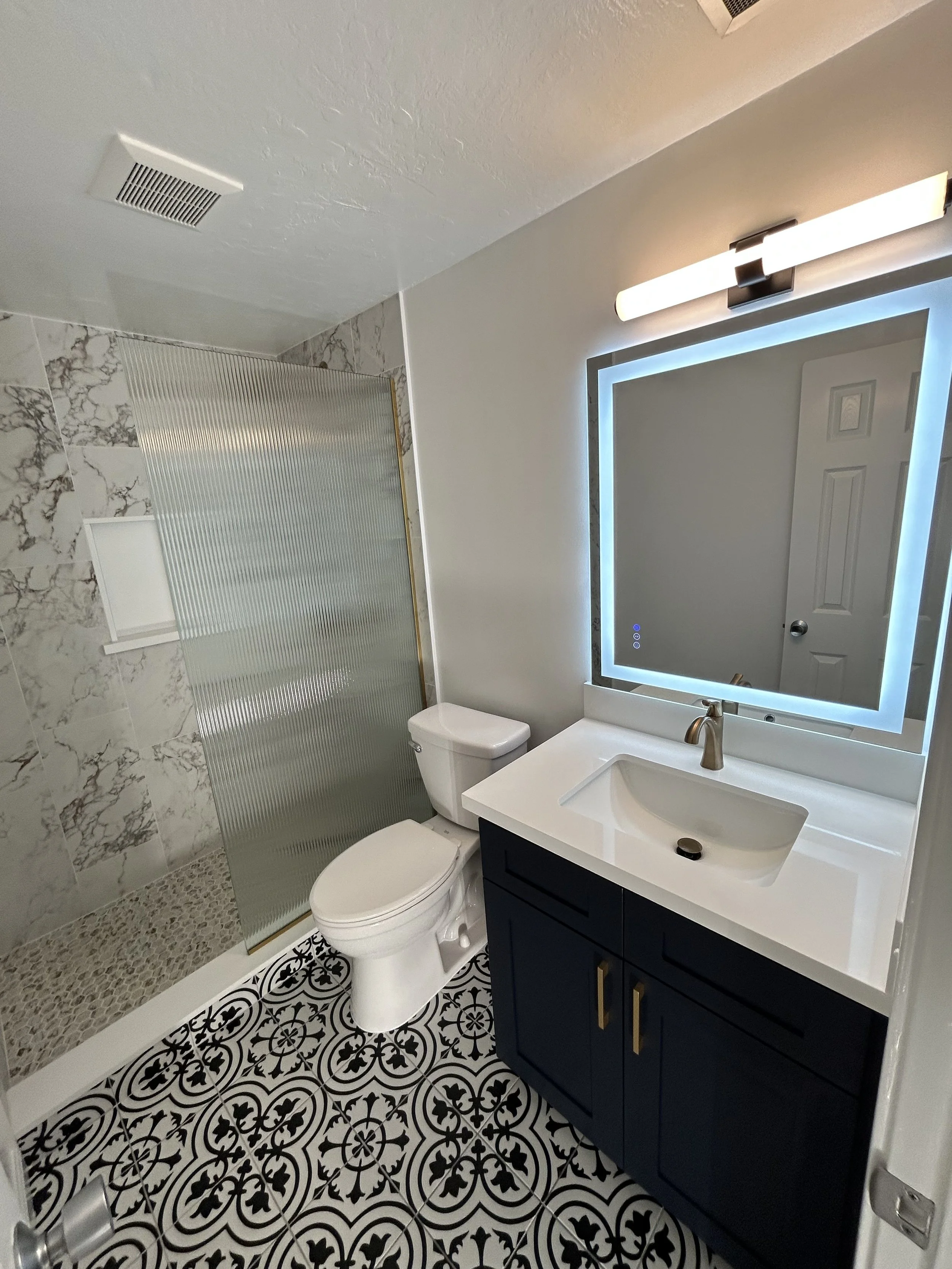 A modern bathroom with a patterned black and white floor, a navy vanity with gold handles, white sink, large backlit mirror, soap dispenser, white toilet, and a glass shower with marble tiled walls and pebble flooring.