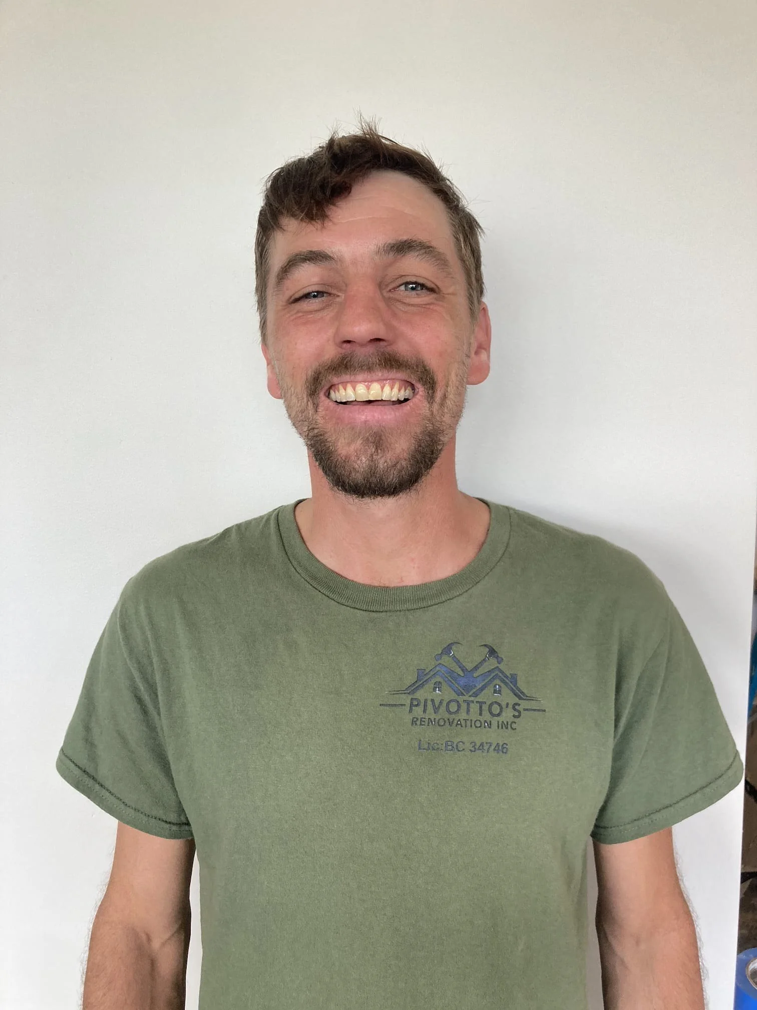 A man smiling with a beard and mustache wearing a green t-shirt, standing in front of a plain white wall.