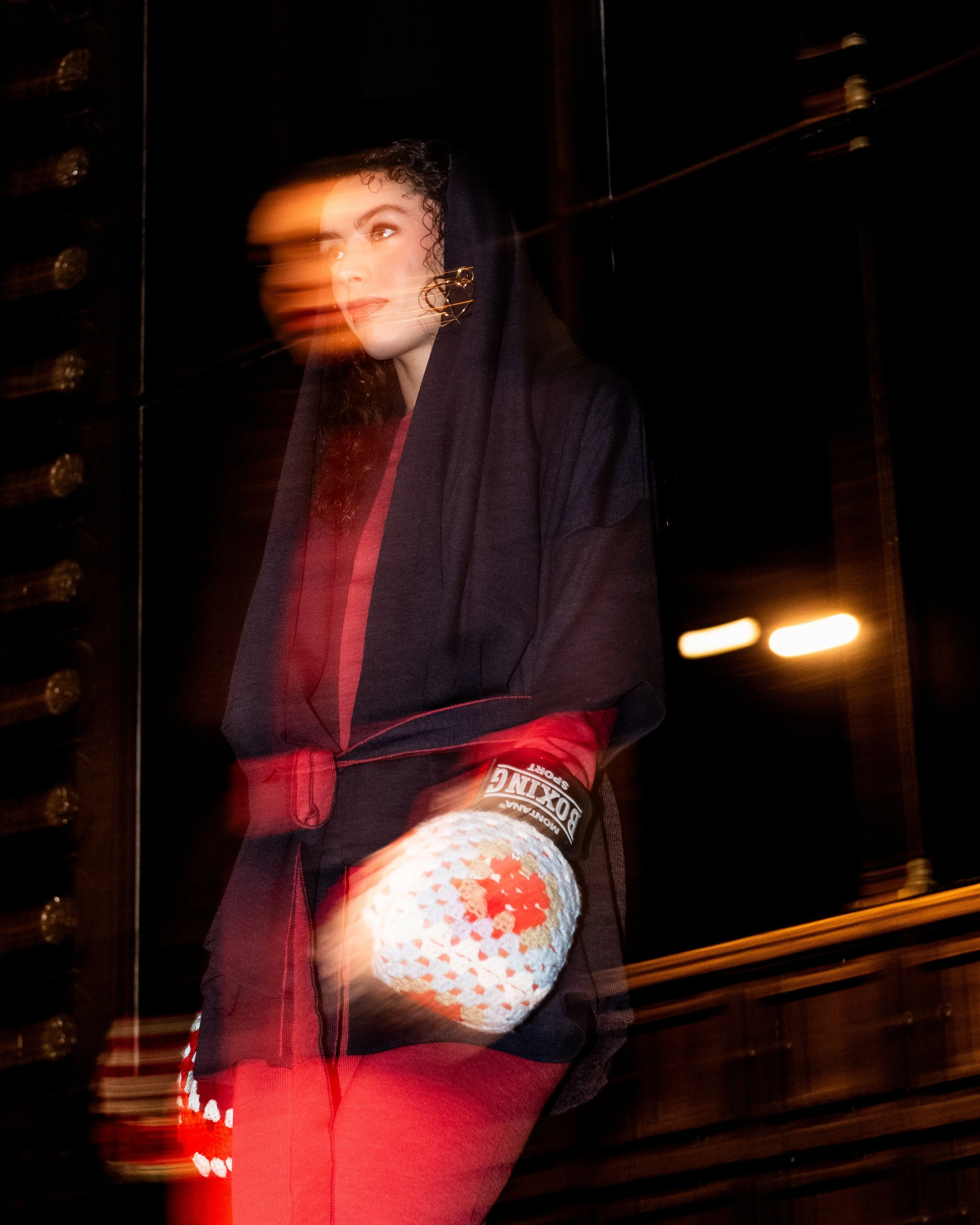 A woman with curly hair wearing a black hooded jacket and gold earrings, standing in a dark room, with a blurred motion effect in the photograph.
