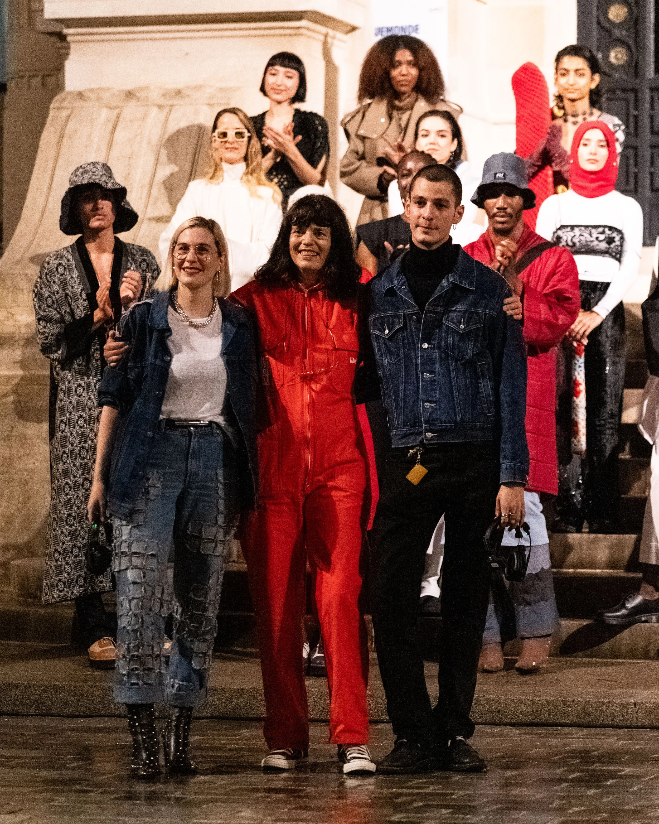 A diverse group of people standing together on a staircase during nighttime, dressed in various stylish and casual outfits, with some clapping and smiling.