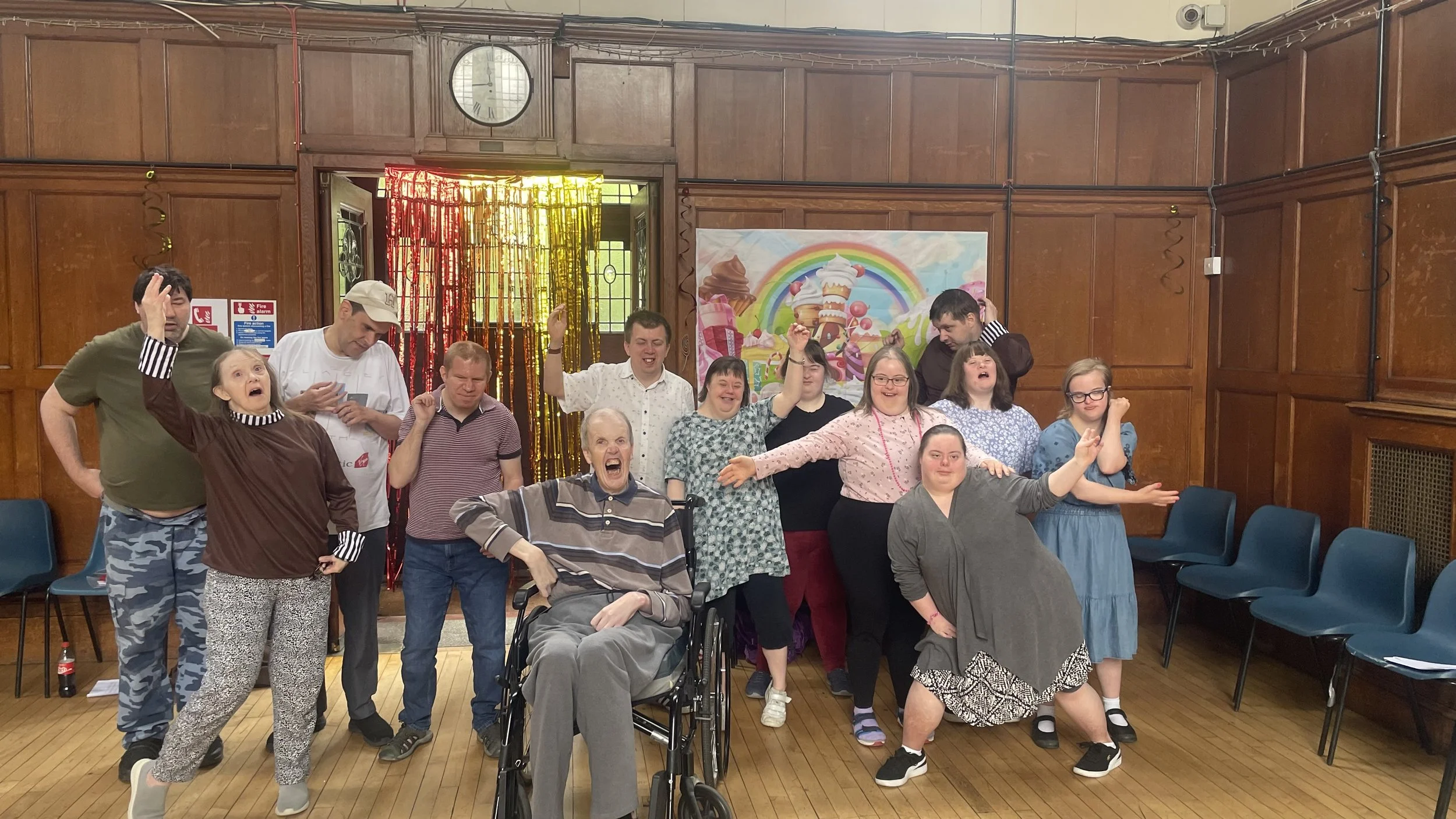 Make a Scene students are a diverse people. Here they are celebrating in a room with wooden walls and a banner with a rainbow and ice cream cones. Many are smiling or cheering, some with arms raised, including one person in a wheelchair.