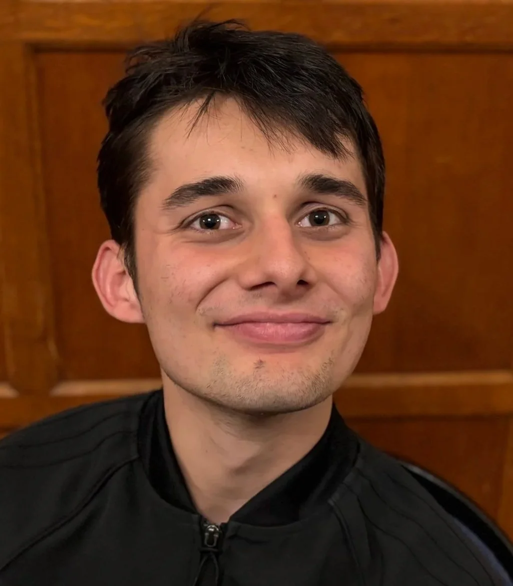 Make a scene student. A young man with dark hair and light skin, smiling, wearing a black zip-up jacket, standing in front of a wooden background.
