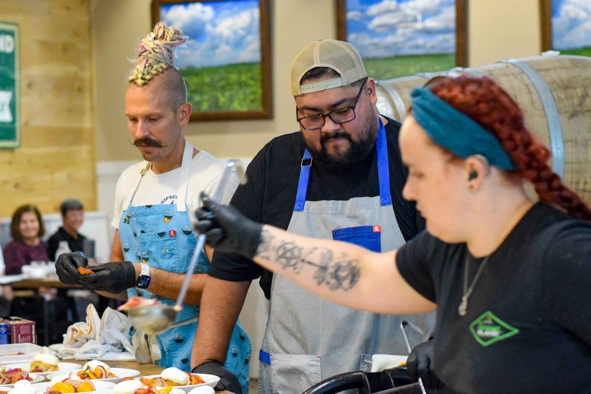 Three people working behind a food counter, preparing dishes with vegetables and other ingredients, at a culinary event or restaurant. Two men and a woman are focused on their tasks.