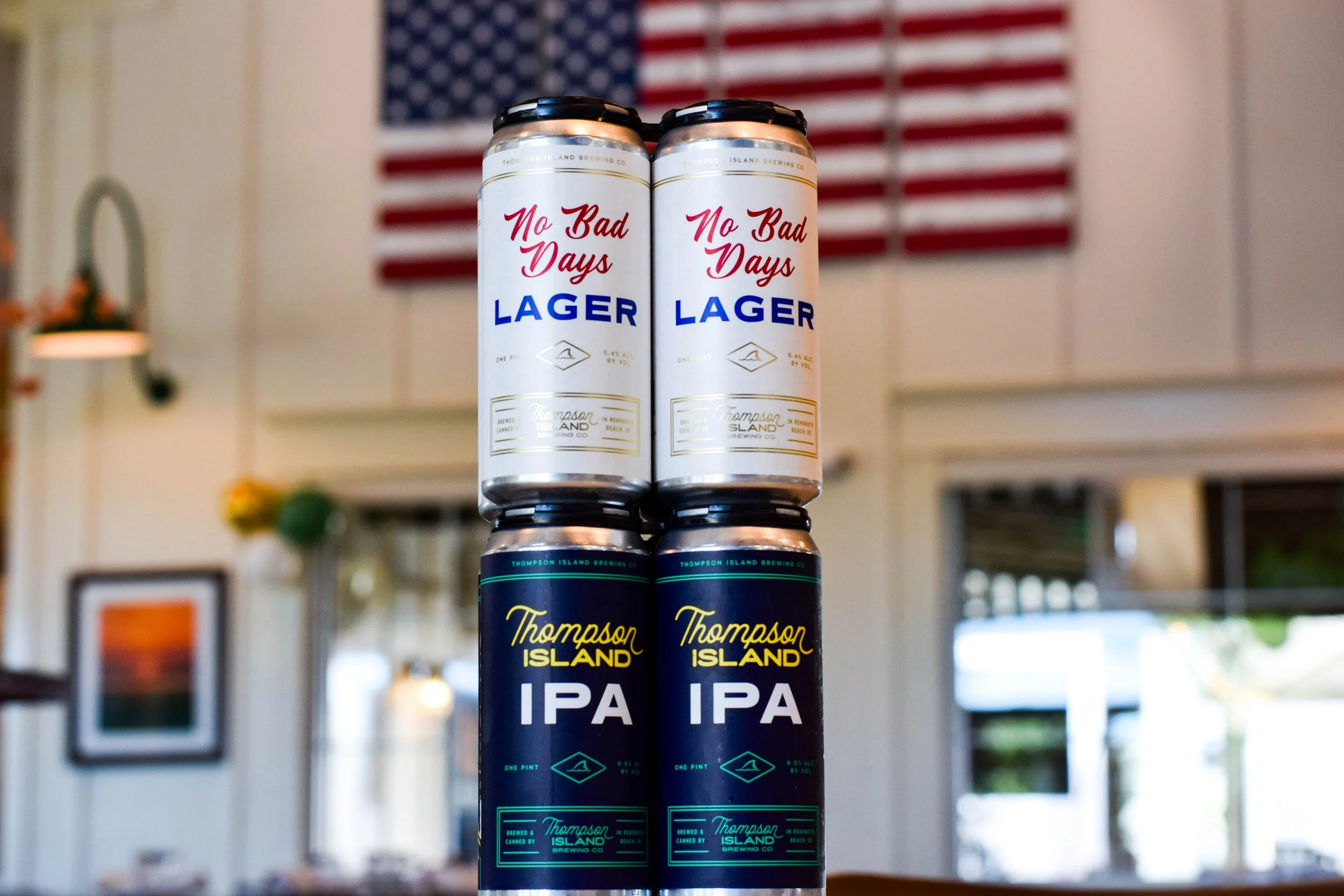 Cans of Thompson Island IPA beer stacked vertically in front of an American flag hanging on a wall.