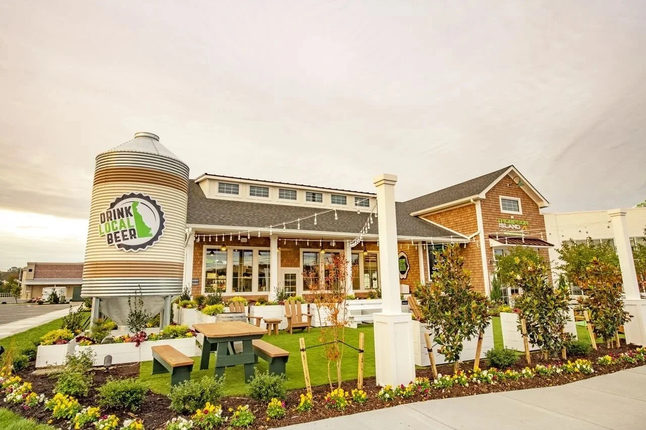 Exterior of a restaurant with a sign that says 'Drink Local Beer', surrounded by landscaped bushes, flowers, and outdoor seating, under a cloudy sky.
