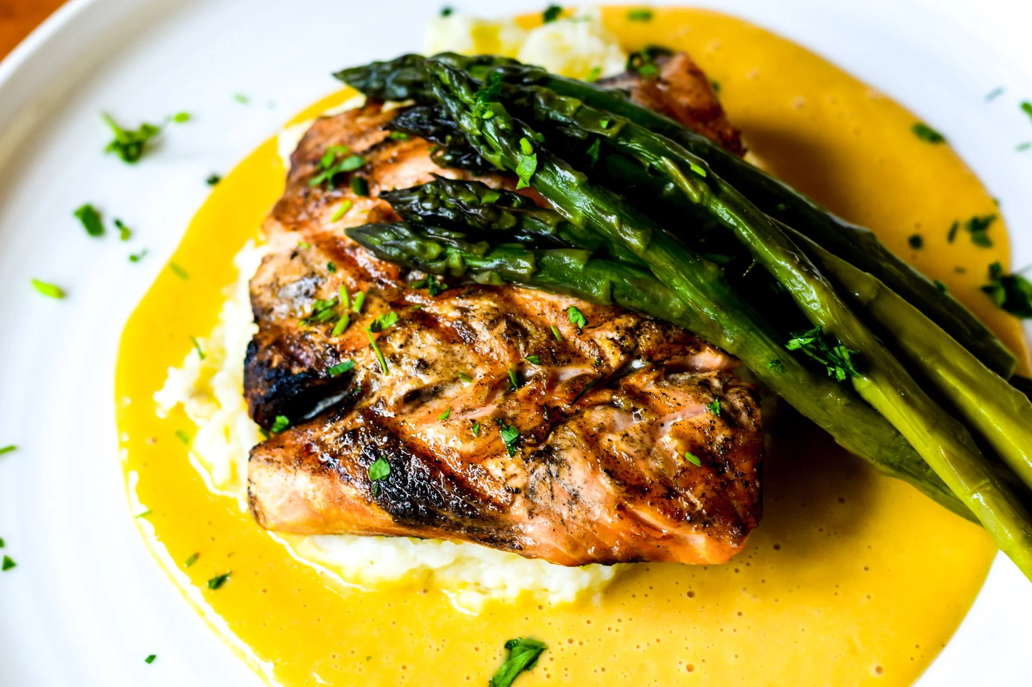 A plated dish featuring a piece of cooked salmon topped with sautéed asparagus, served with mashed potatoes and a yellow sauce, garnished with chopped herbs.