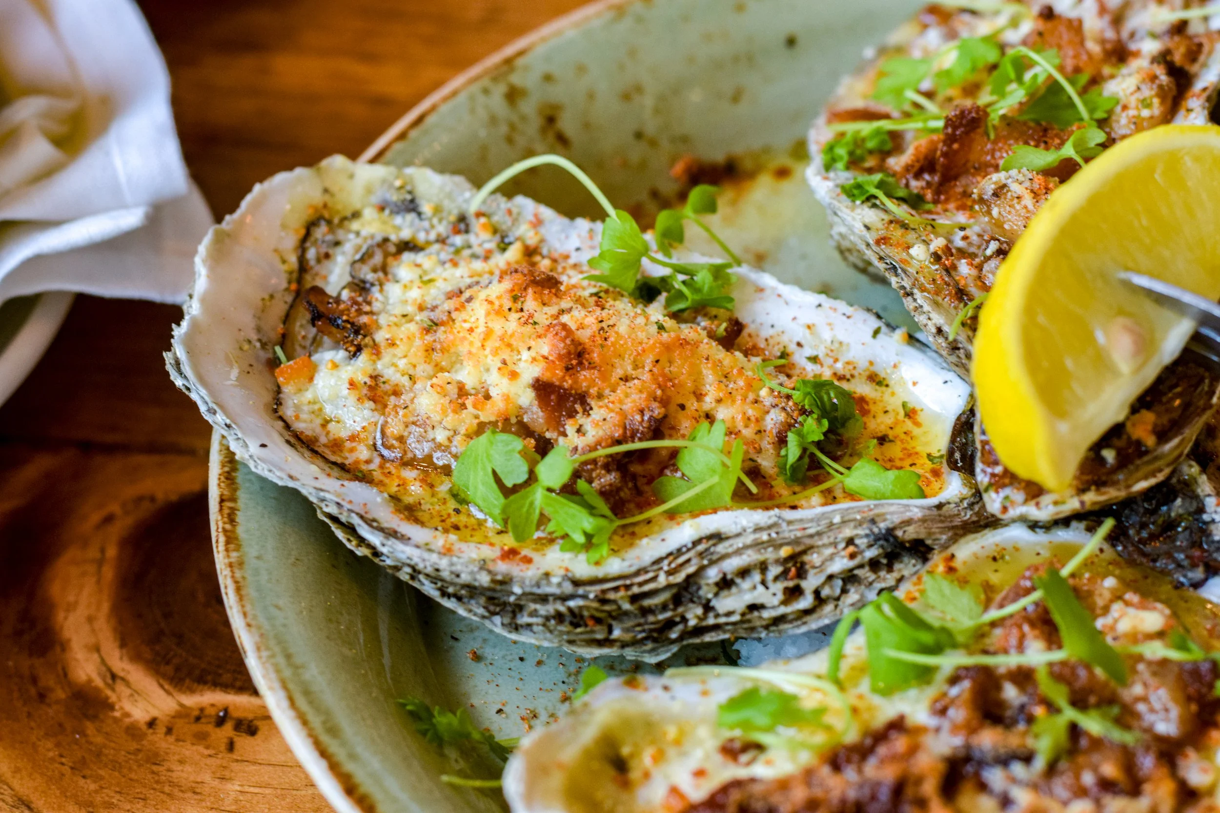 A close-up view of baked oysters topped with herbs, seasoning, and sauce, garnished with fresh green microgreens and a lemon wedge, served on a ceramic plate.