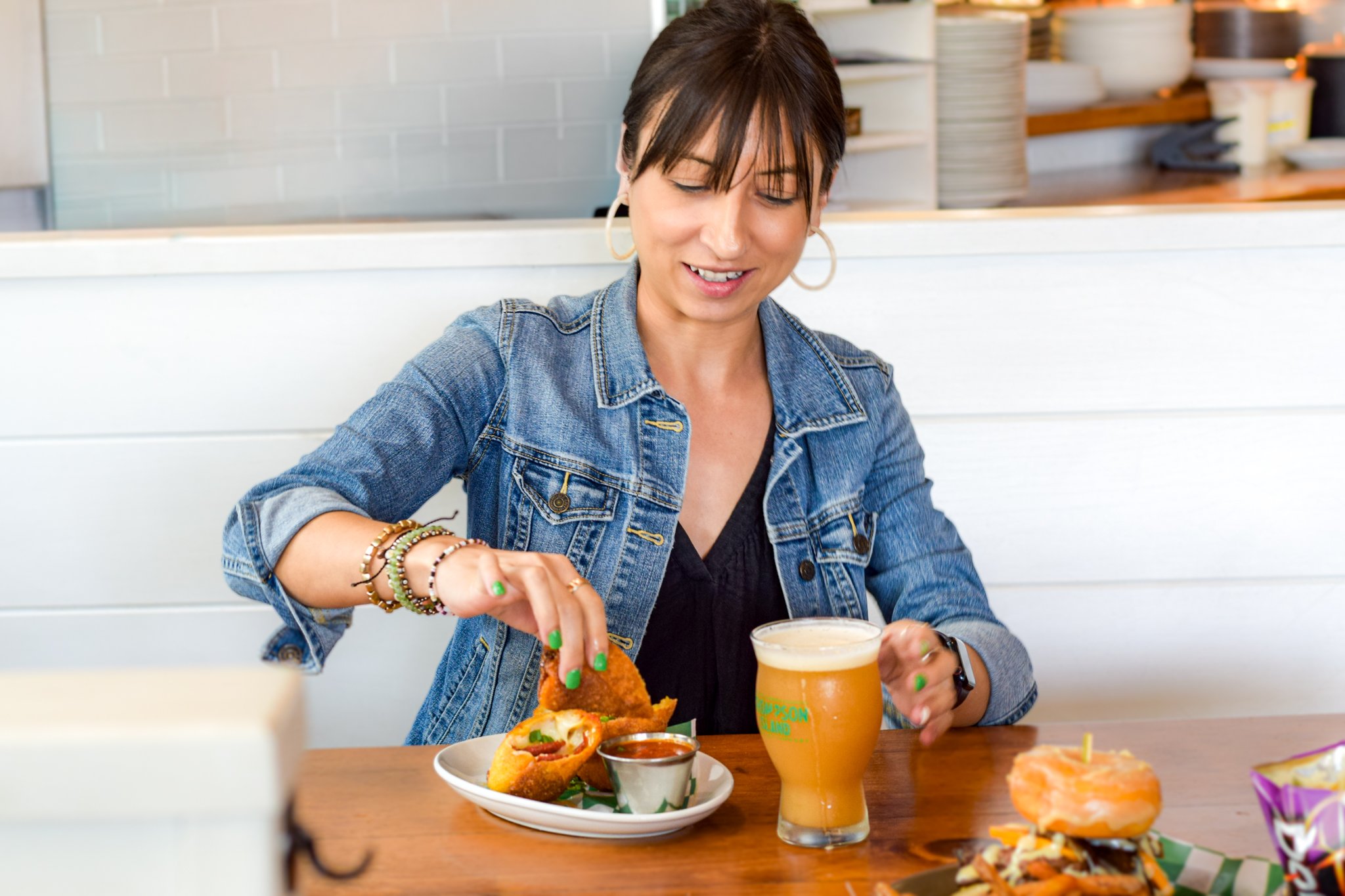 A woman with short dark hair wearing a denim jacket and jewelry is smiling and enjoying a meal at a restaurant table. She has a plate of tacos, a glass of beer, and various colorful dishes on the table.