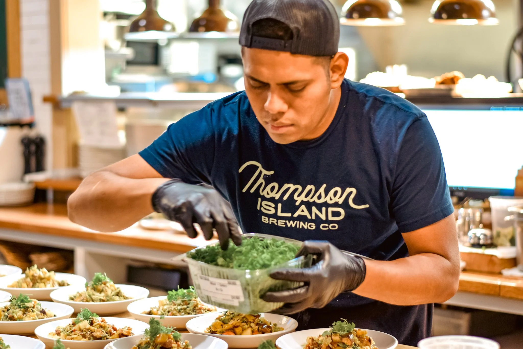 A man wearing a black Thompson Island Brewing Co t-shirt and a backward gray cap is serving food at a buffet with bowls of salad.