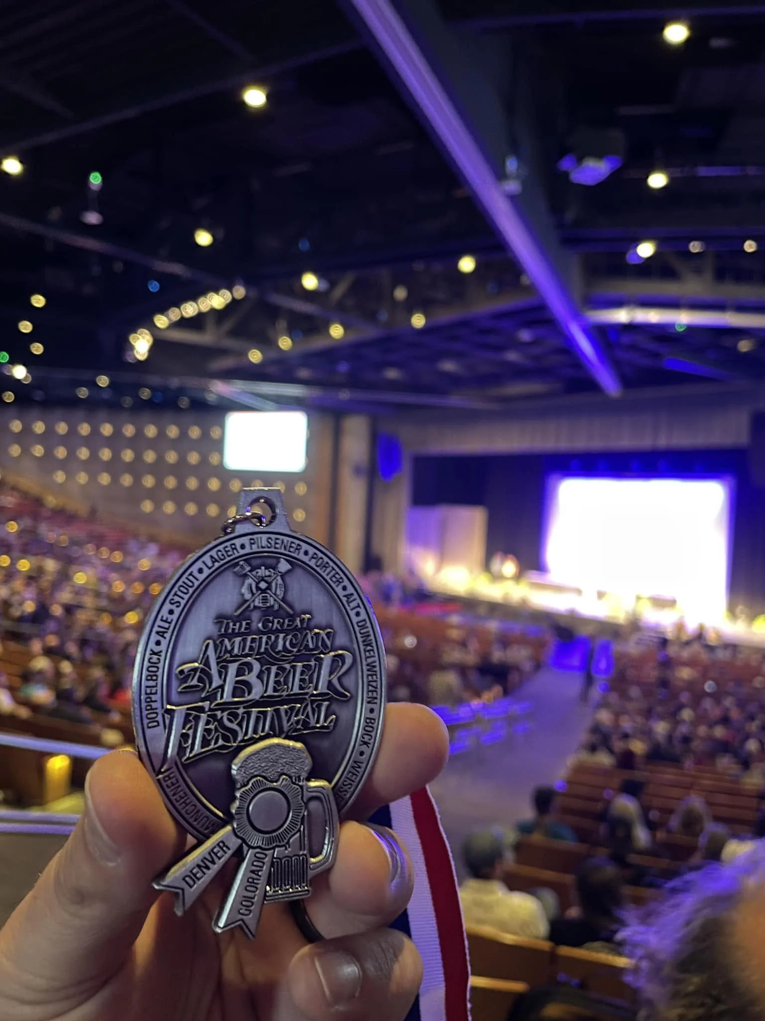 Person holding a silver medal with a cityscape and festival text in the foreground, seated inside a dimly lit auditorium or concert hall with stage lighting and audience seating visible in the background.
