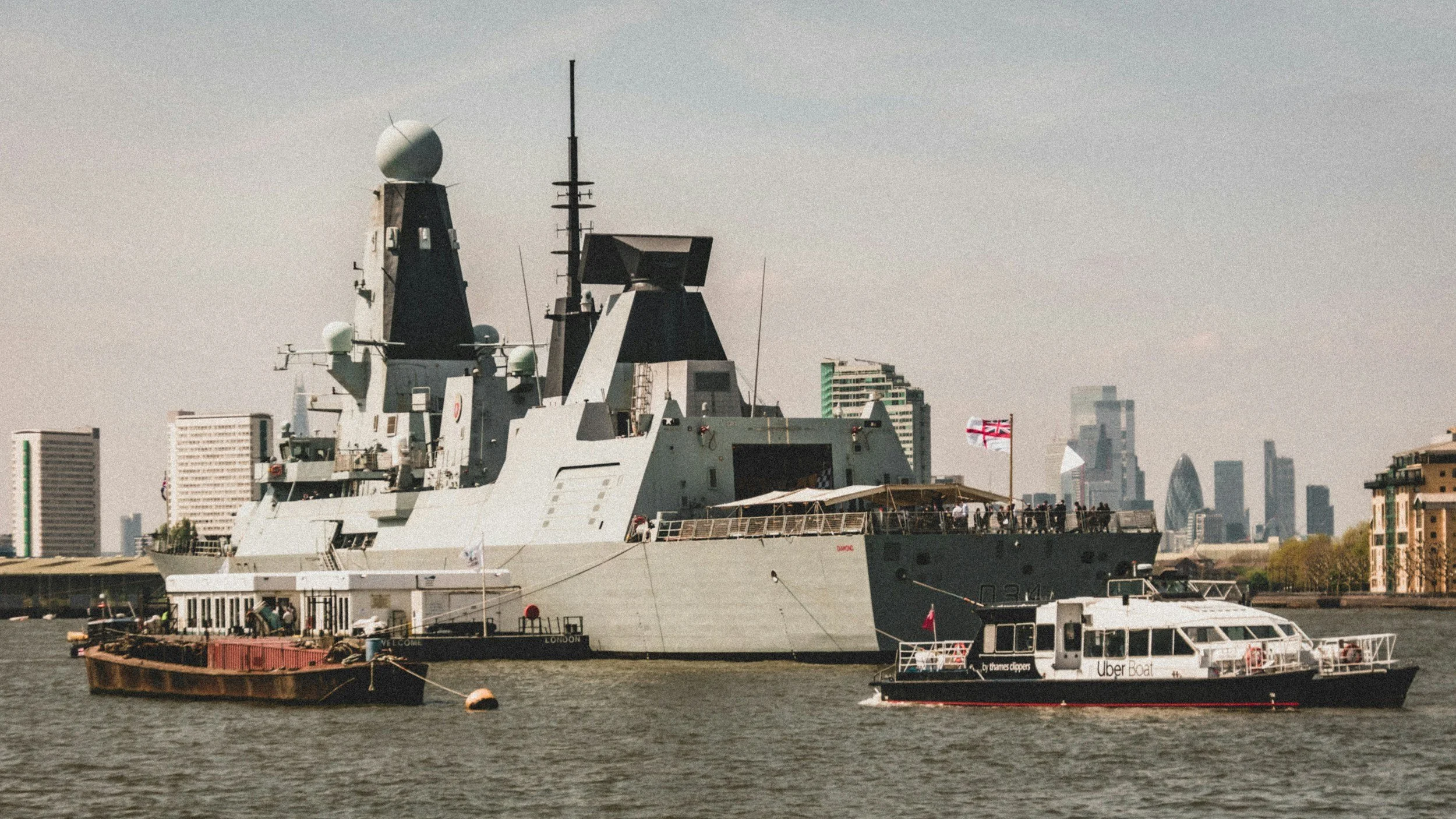 A large gray naval ship with modern architecture on the river, with London city skyline in the background, including the Gherkin building, and an Uber Boat Thames Clipper docked nearby.