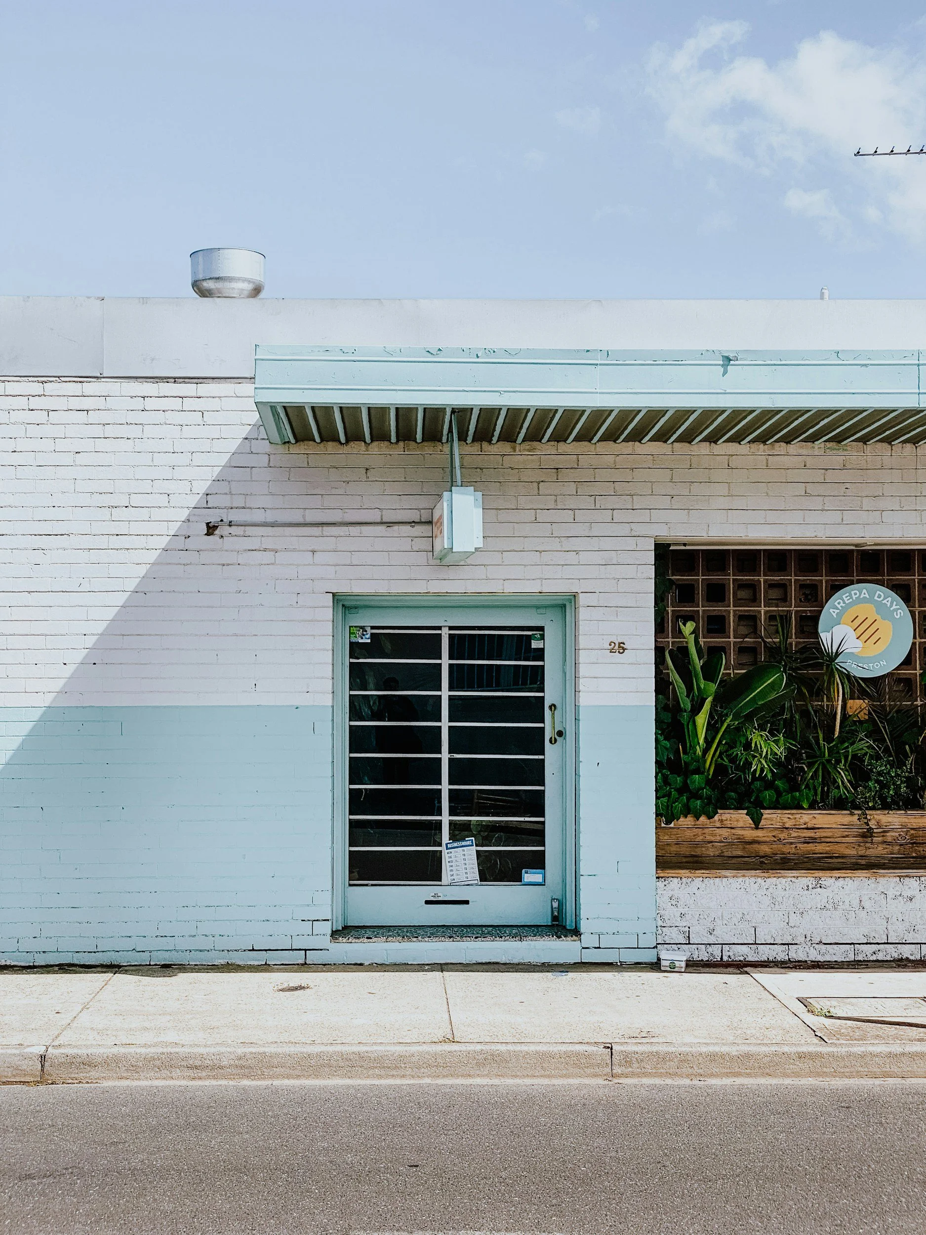 Front of a white brick building with a glass door, plants in a window planter, and a sign that reads "Arepa Days" on the right side.