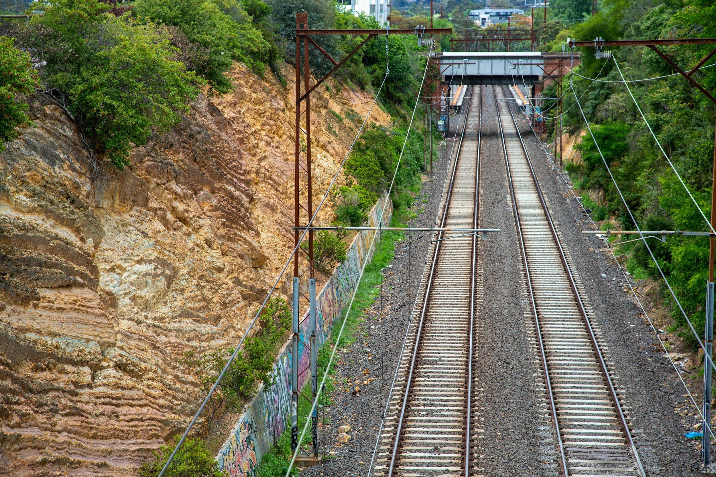 View of train tracks surrounded by green trees on a hillside, with a graffitied wall on the left and a bridge in the distance.