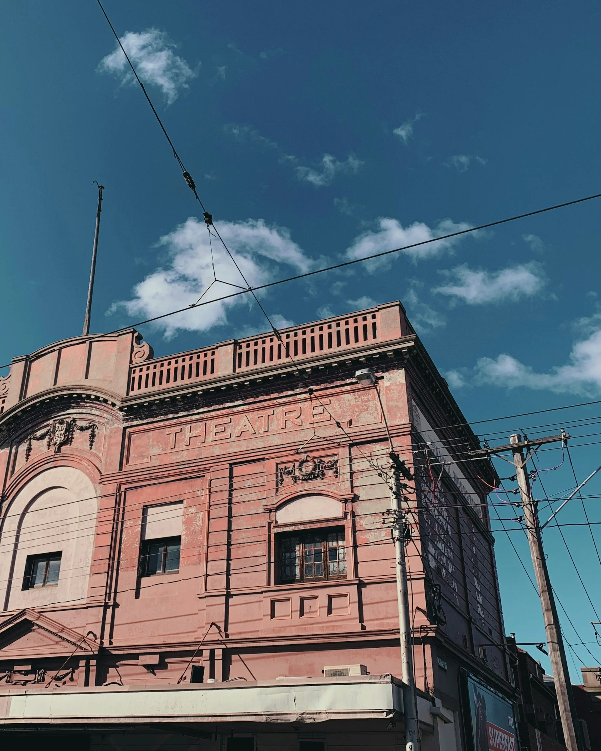 An old pink theater building with the word 'THEATRE' inscribed on the upper facade, with windows, decorative details, and overhead wires against a blue sky with scattered clouds.