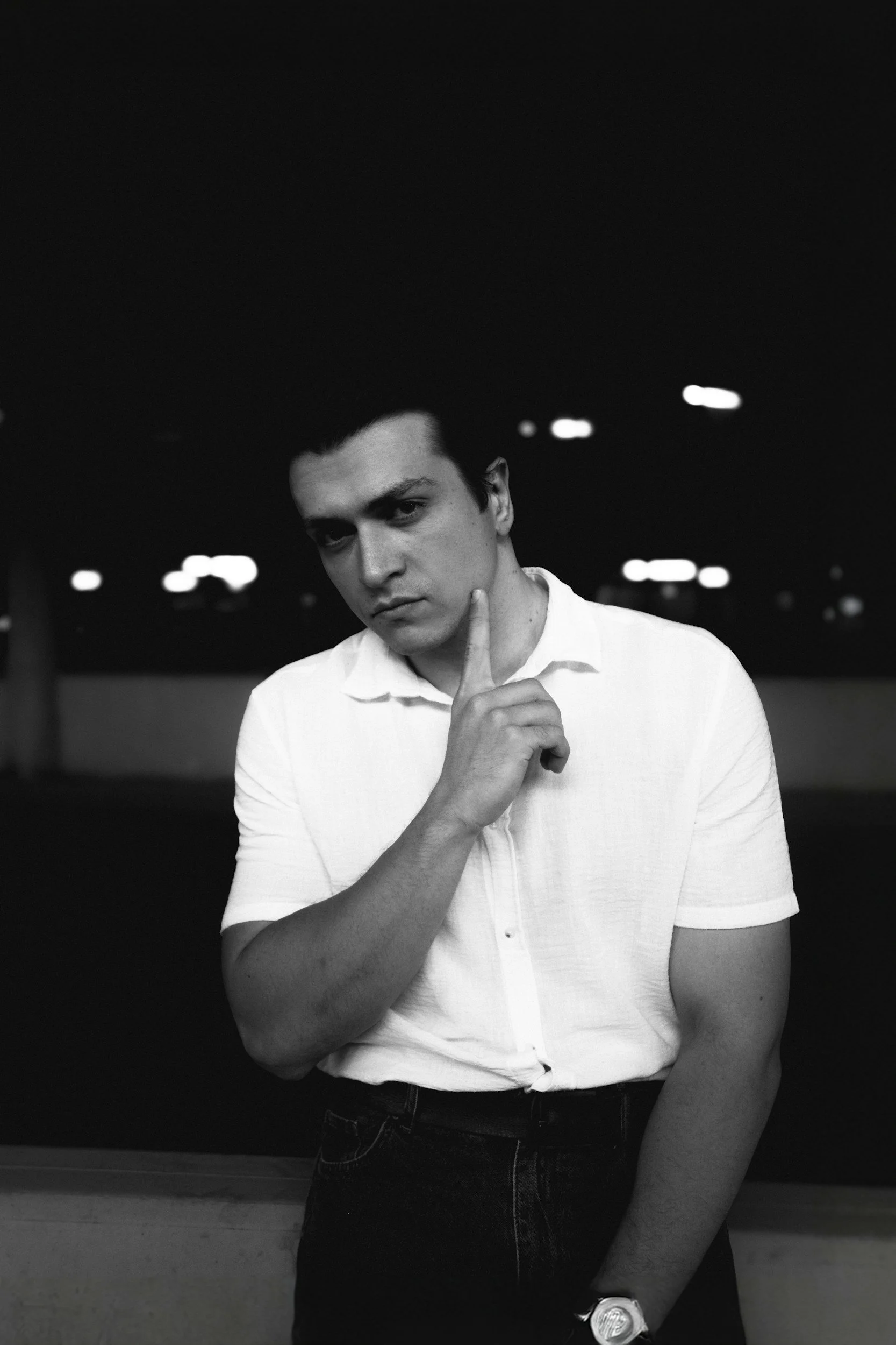 A young man with dark hair, dressed in a white short-sleeve shirt and black pants, stands indoors at night, looking serious while holding his right index finger to his cheek. The background is dark with some small lights visible.