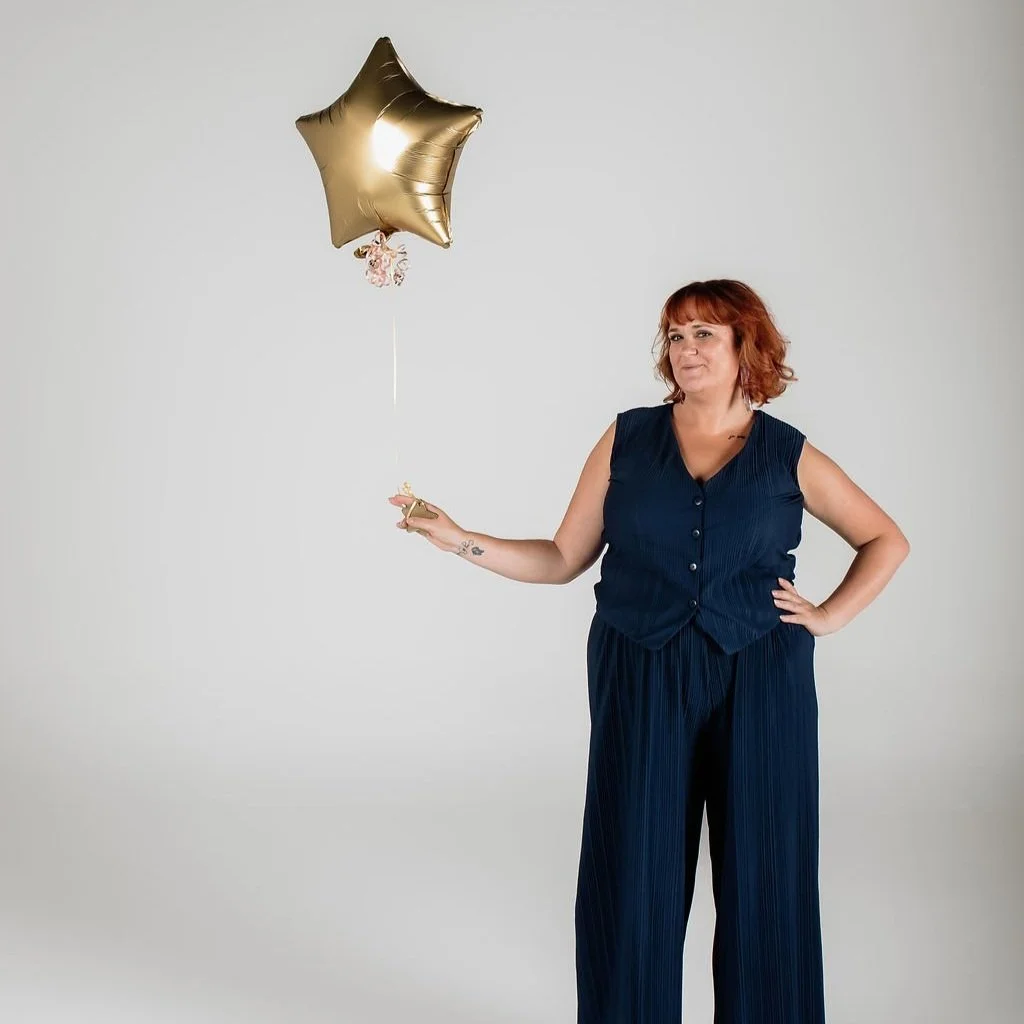 A woman with red hair wearing a navy blue sleeveless top and wide-leg pants, holding a gold star-shaped balloon in her right hand against a plain light gray background.