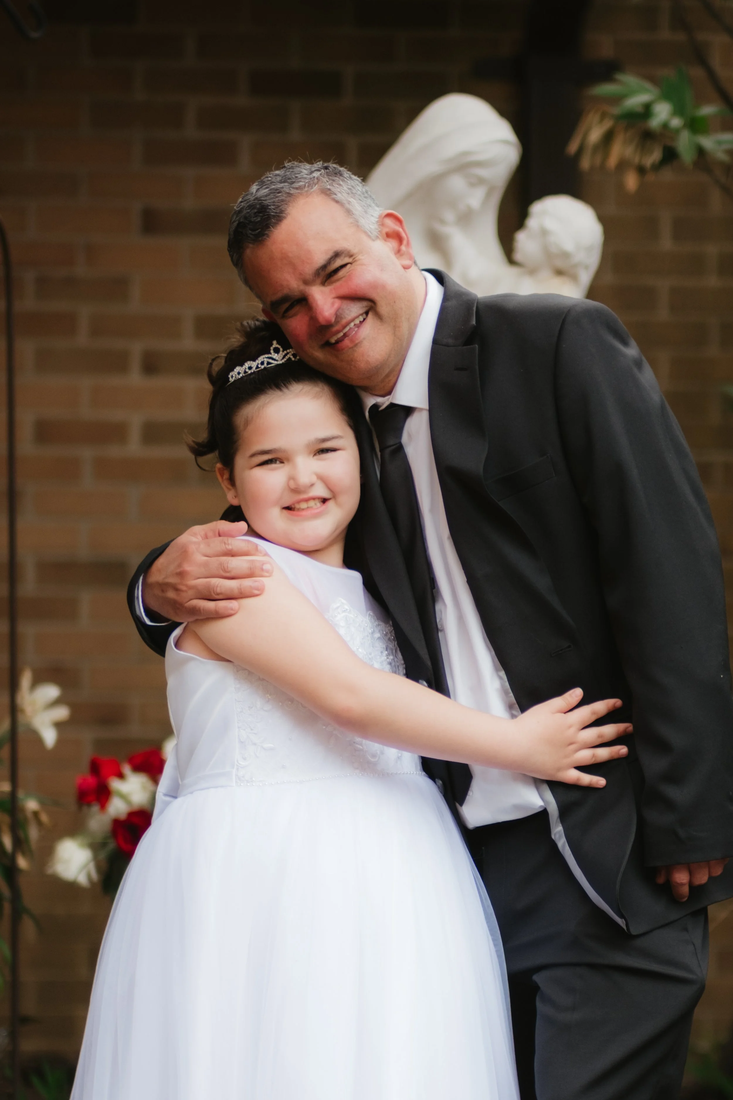 A young girl in a white dress and tiara hugging an older man in a black suit and tie, both smiling at the camera, with a religious statue and brick wall in the background.
