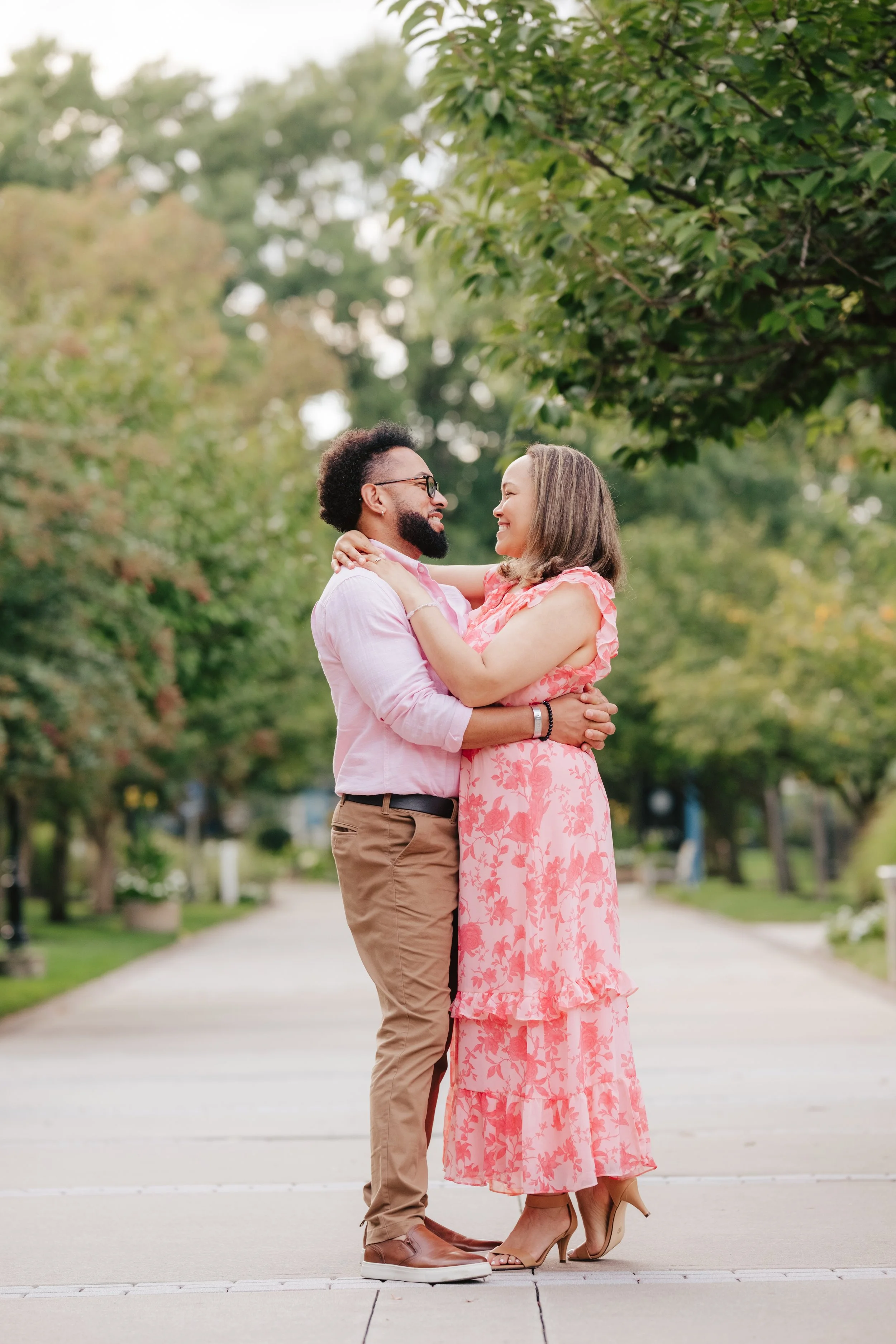 A couple standing on a park pathway, embracing each other and smiling while looking into each other's eyes, with trees in the background.