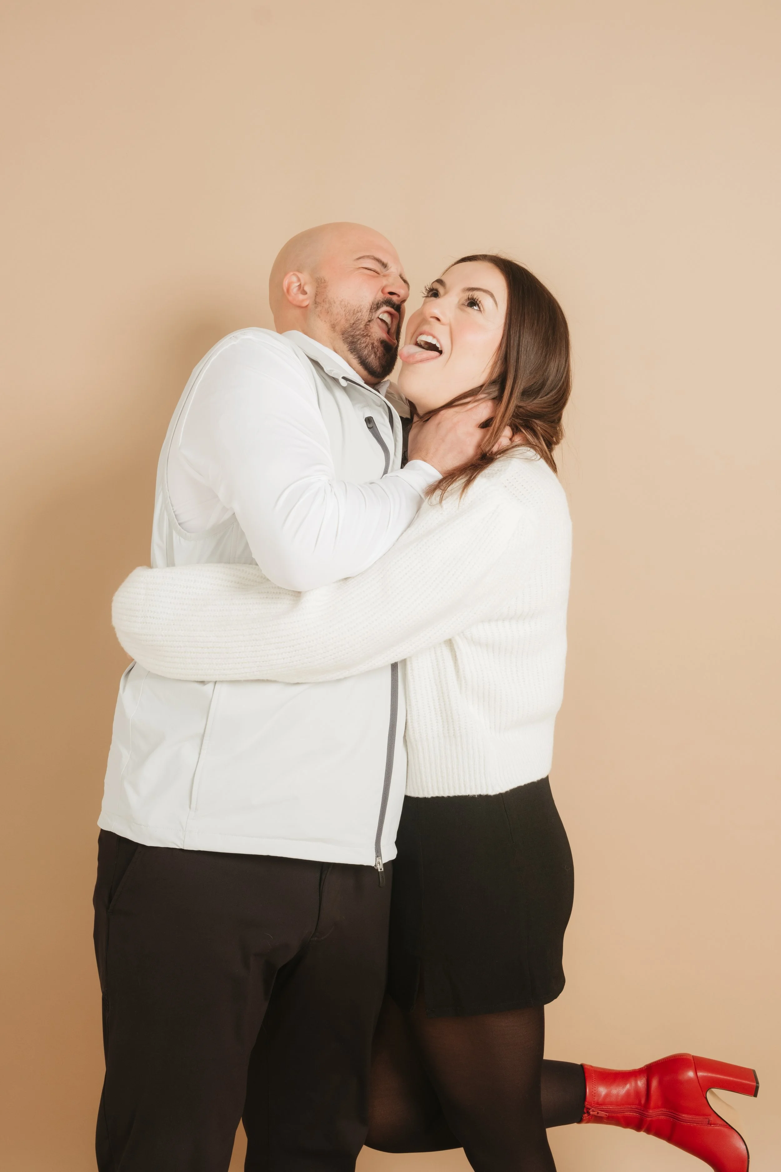 A man and a woman playfully fighting or joking, with the man holding the woman's neck while she sticks out her tongue and lifts her leg, wearing red boots.