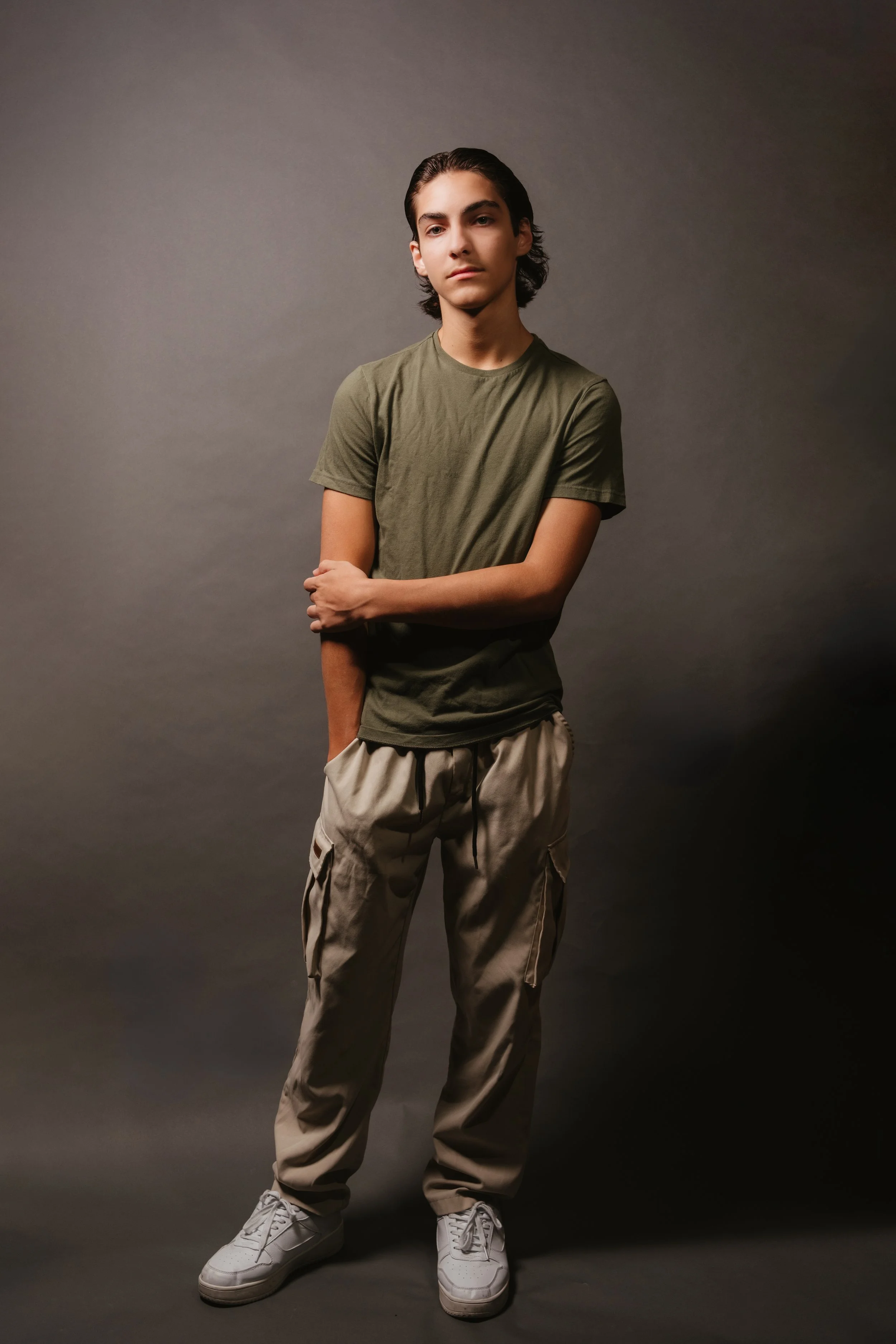 A young man with dark hair wearing a green t-shirt, beige cargo pants, and white sneakers against a plain gray background.
