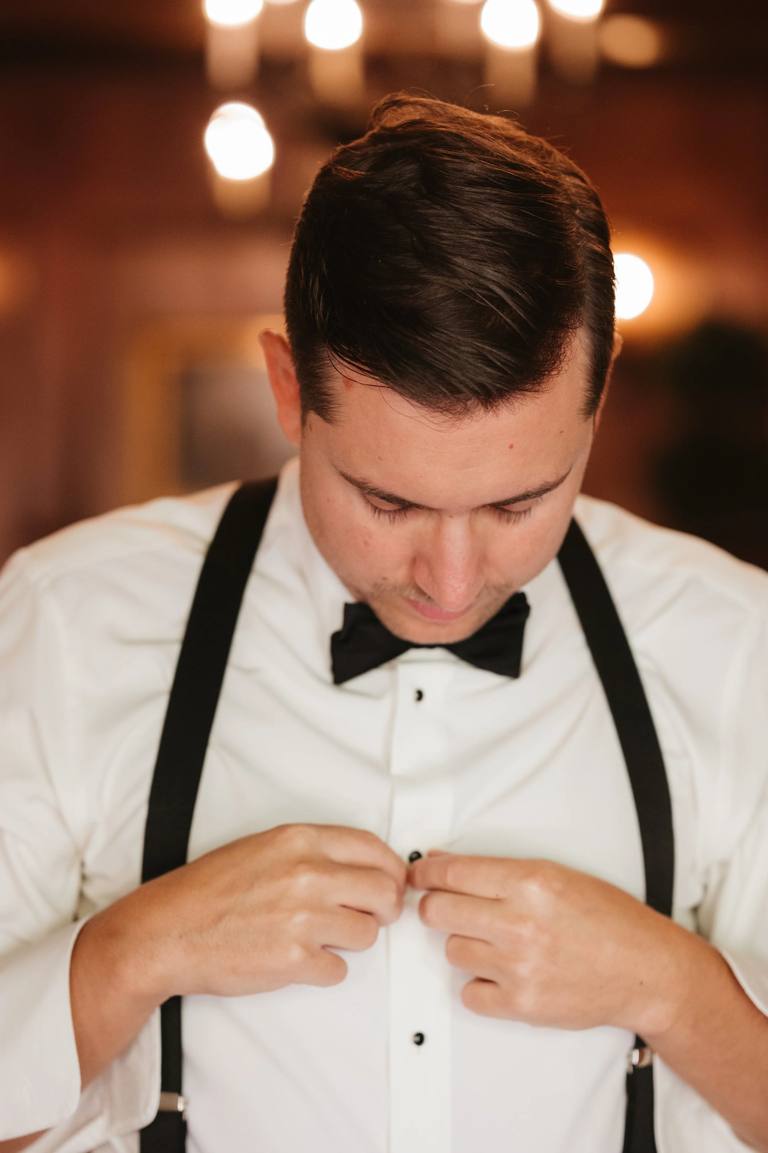 A man dressed in a white shirt and black bow tie, adjusting his buttons, with suspenders over his shoulders in a warmly lit setting.