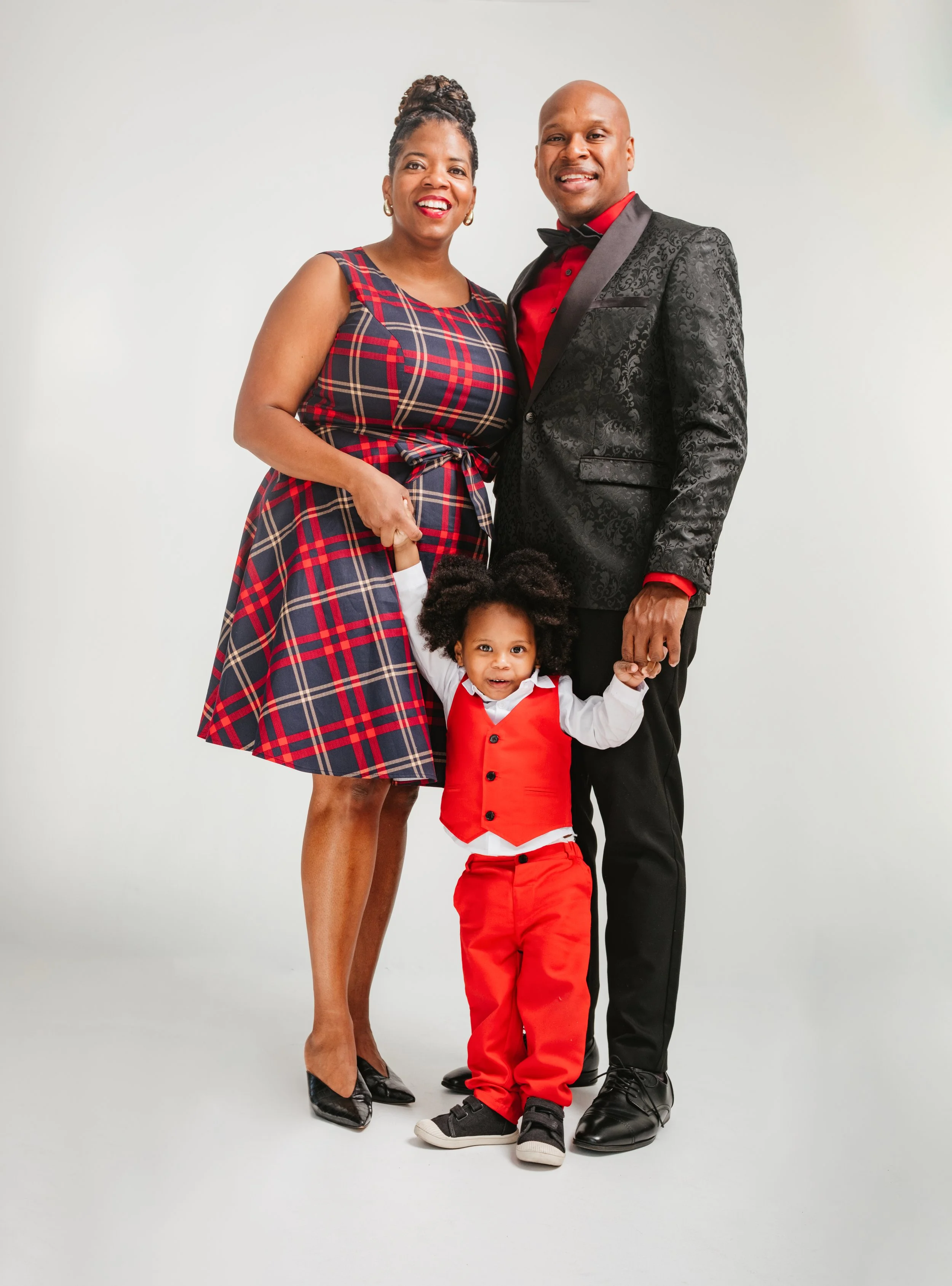 A family of three dressed in formal attire, standing together against a white background. The woman is wearing a plaid dress, the man is in a black tuxedo with a red shirt, and the child is in a red vest and pants with a white shirt.
