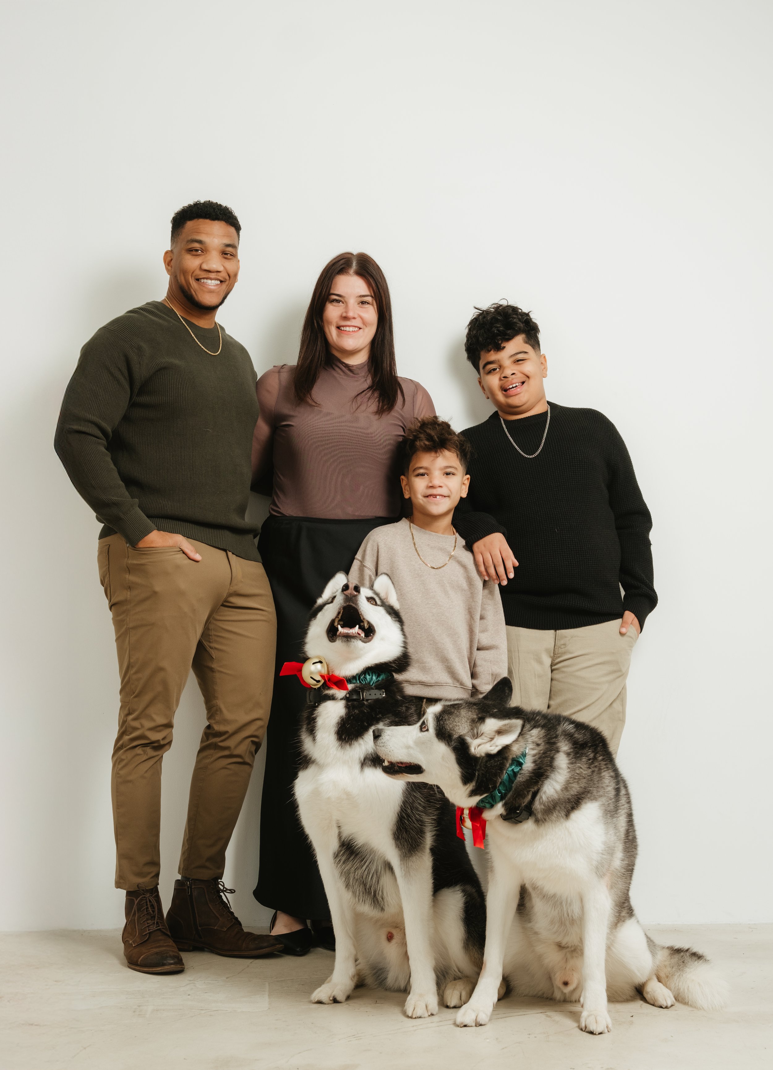 Family portrait of two adults, two children, and two Siberian Huskies posing together against a white wall.